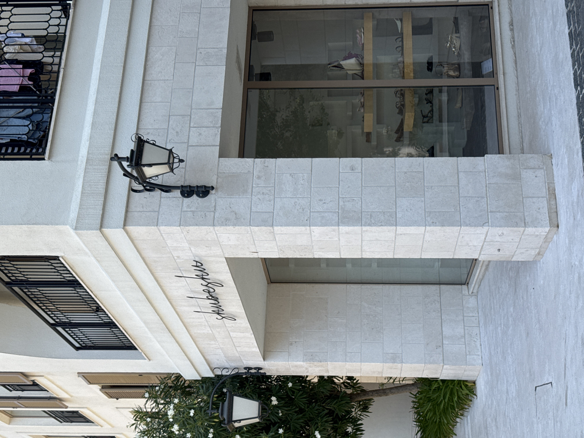 A storefront displays shoes in Tivat, Montenegro. The shop's name, "shubeshu," is written in cursive on the light-colored stone facade. A large window reveals shelves stocked with various shoes and handbags. A black wrought-iron lamppost is affixed to the building's exterior. In front of the store, a leafy green tree with white flowers stands next to lush green foliage. The scene is set against a backdrop of cream-colored, brick-like stonework and a clear, light-colored sky.