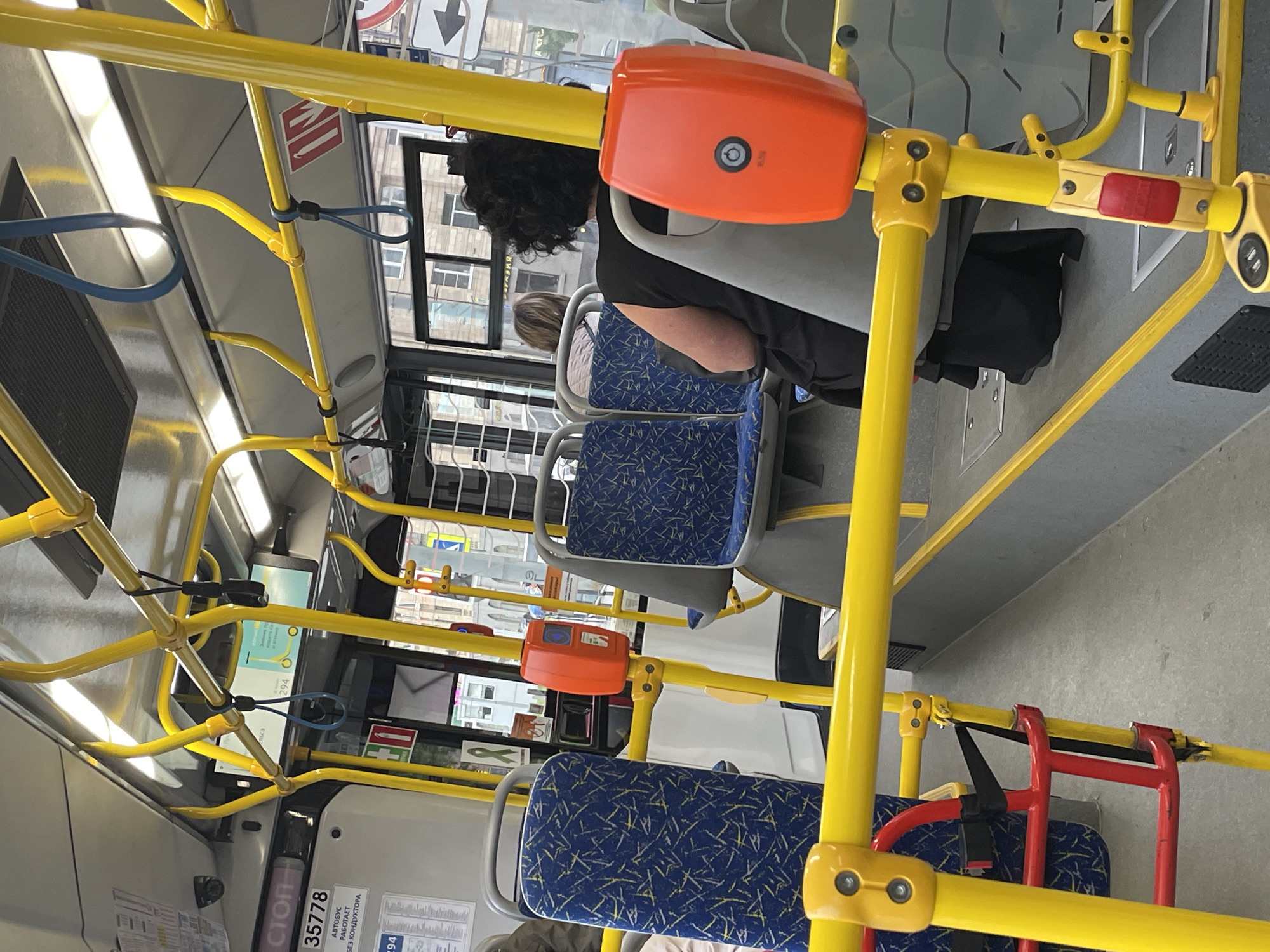 The image captures the interior of a public transport vehicle, likely a bus or tram, in Saint Petersburg, Russia. The dominant color scheme is bright yellow, used for the handrails and structural supports, contrasting with the blue patterned seats and gray flooring. Several passengers are visible, including a woman with dark curly hair seated in the foreground, facing away from the camera and wearing a black dress. Another passenger with lighter hair is seated further back, also facing away. Various fixtures are present, such as orange ticket validators mounted on the yellow poles, and a red and black child safety seat structure on the left. The overall environment suggests an urban transportation setting, with glimpses of buildings through the windows indicating a city street outside. The lighting appears to be a combination of artificial overhead lights and natural daylight filtering in.