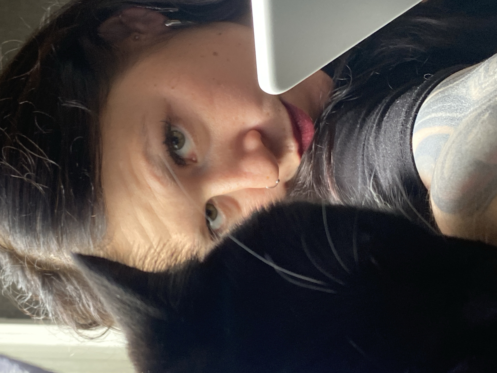 The media file is a close-up, overhead shot of a person and a black cat, with a white object partially visible in the upper right corner. The person appears to be looking down, with their face tilted. Their eyes are green, and they have dark hair, freckles, and a nose ring. Their lips are red. The cat is black and largely fills the bottom of the frame, obscuring much of the person's body. The white object appears to be a flat surface, possibly a laptop or tablet, positioned above and to the right of the person. The lighting suggests an indoor setting, possibly with artificial light. The overall mood is intimate and candid, with the person and the cat seemingly interacting or sharing a moment. There are no visible text elements in the image. The location context of Chaikovsky, Russia, does not provide any further visual cues within the image itself.