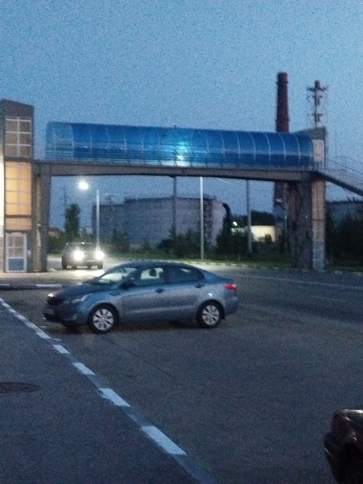 The image depicts an outdoor scene at dusk, possibly in an industrial or commercial area of Kostanay, Kazakhstan. A blue, enclosed pedestrian bridge spans across the scene, supported by concrete pillars. To the left, a building with large windows and lighting is visible. In the foreground, a silver sedan is driving on an asphalt road marked with white lines. Further in the background, another car with headlights on is stationary. A tall, brick industrial chimney is visible against the dusky sky, along with some trees and other industrial structures. The overall atmosphere is subdued due to the low light conditions.