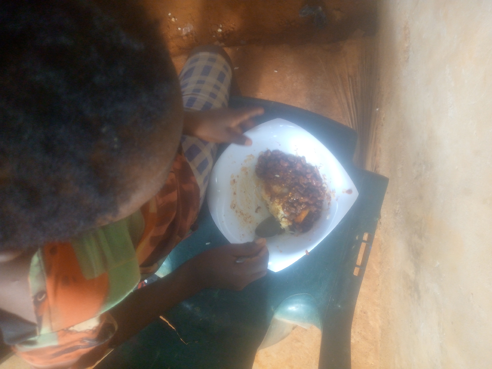 An overhead, high-angle shot captures a person eating a meal. The individual's head and shoulders are visible from behind, showing dark hair and skin. They are seated, with a white, square plate placed on a dark green plastic chair or stool in front of them. The person holds a spoon in their right hand, which is resting in the food on the plate.
The meal consists of a base of light-colored grains, likely rice, topped with a generous portion of a dark reddish-brown, chunky stew or sauce. Some food particles are scattered on the plate and the surrounding area.
The person is wearing clothing that includes a plaid pattern in white and purple on their lower half, and a shirt or top with green and orange sections on their upper body. The setting appears modest, with a reddish-brown floor, possibly earth or unpolished concrete, and a plain light beige wall in the background. The lighting suggests daytime. There is no visible text or identifiable location other than a simple indoor or sheltered outdoor space.