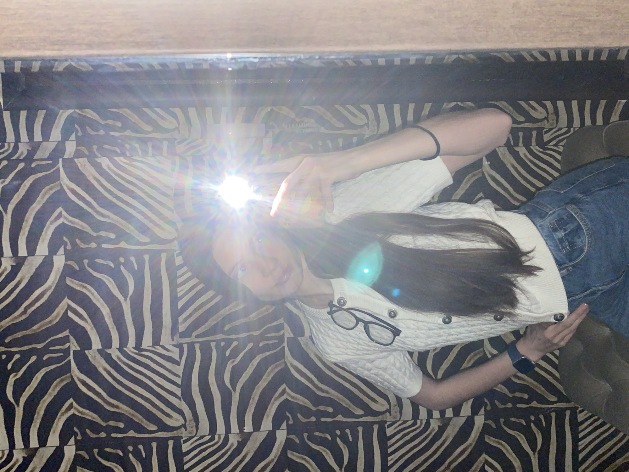 A selfie captures a young person lying horizontally on a surface with a zebra-print pattern, likely a floor or large rug, indoors. A bright camera flash emanates from the upper-left of the image, near the subject's face and left hand, where a smartphone is being held to take the picture. The flash obscures some facial details, but the person appears to have long, dark hair and is looking directly towards the camera.
The subject is dressed in a white, cable-knit cardigan with dark, possibly silver-toned, buttons running down the front. A pair of black-rimmed eyeglasses rests on the cardigan near their right shoulder. They are also wearing blue denim jeans, visible towards the right side of the frame, and a dark-colored smartwatch on their left wrist.
The background consists of a distinctive zebra-stripe pattern in dark brown/black and light beige/grey, arranged in rectangular sections. Above the subject's head, a solid, light-colored surface, possibly a wall or ceiling, is visible. The strong flash suggests the photograph was taken in a dimly lit environment or for a specific photographic effect. The image conveys a casual, personal moment. No visible text is present. This scene is located in Votkinsk, Russia.