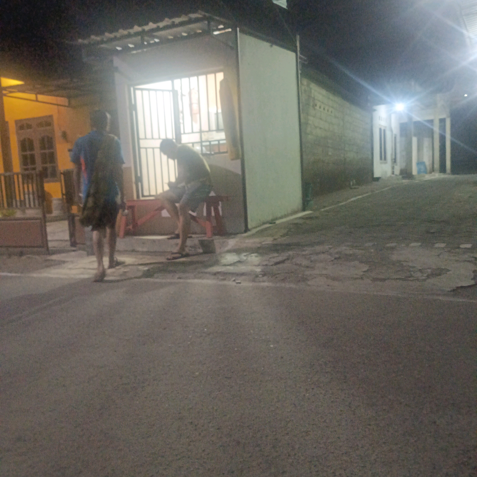 The image shows a dimly lit outdoor scene, likely at night, in Salatiga, Indonesia. In the foreground, a paved road with visible cracks and markings stretches across the frame. To the left, a building with yellow walls and a covered overhang is partially visible, featuring a window with dark bars. Adjacent to it is a white structure with a metal gate.
Two individuals are present in the middle ground. On the left, a person wearing a blue shirt and a backpack is walking away from the camera, their back mostly to the viewer. They appear to be barefoot. To the right, another person is seated on a red bench in front of the gated opening. This individual is wearing a dark shirt and shorts.
The background includes a concrete block wall and a building with multiple windows and vertical supports, illuminated by a bright light source. The overall atmosphere is nocturnal and somewhat stark, with the lighting casting long shadows and creating a grainy effect on the image.