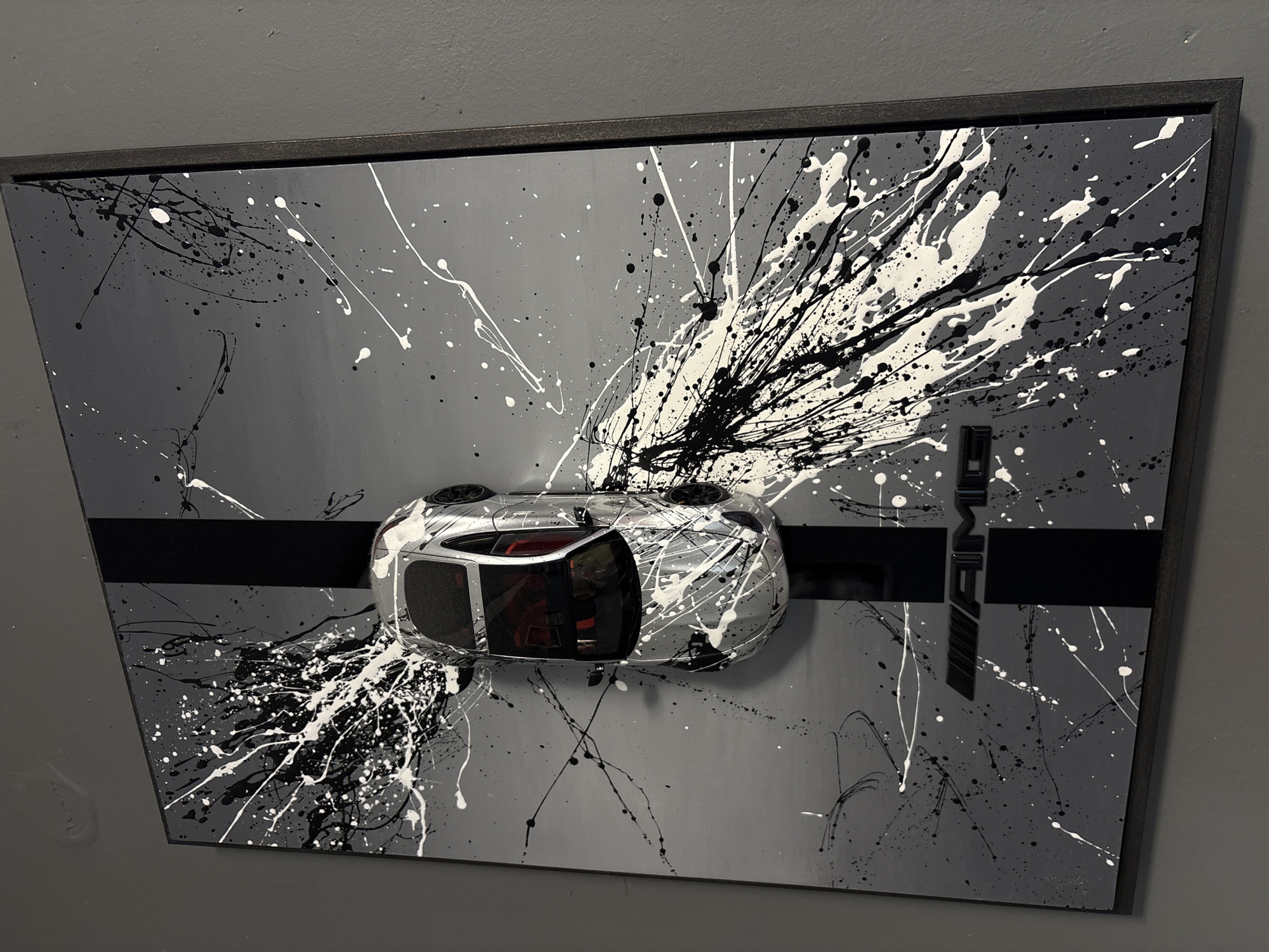 A framed piece of art hangs on a gray wall. The art features a gray background with black and white paint splatters, a black horizontal stripe, and the letters "AMG" in the upper right. A silver model car is affixed to the canvas, positioned on the stripe. The car appears to be a hatchback with a red interior. The scene appears to be indoors.