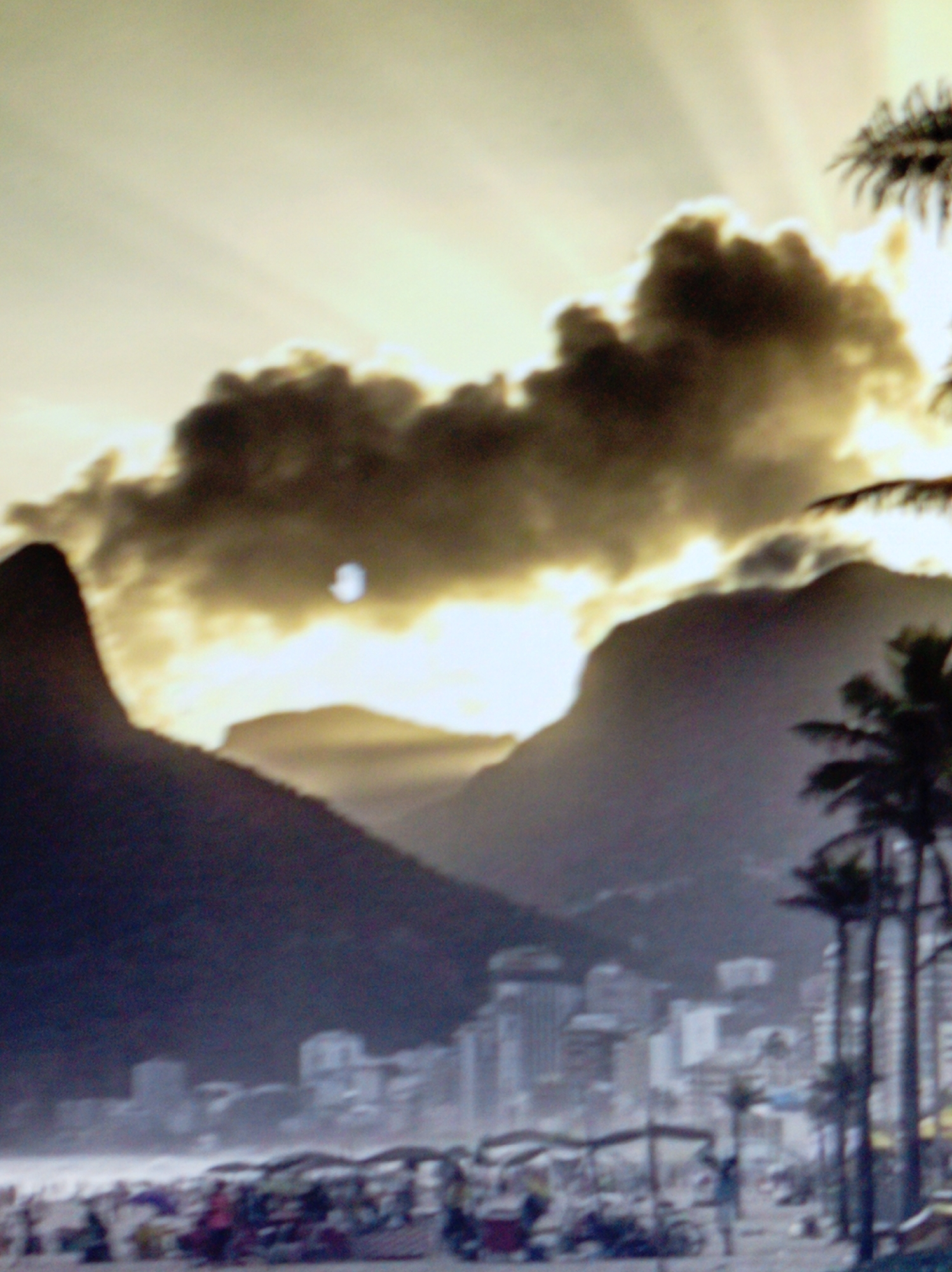 This is a blurry, atmospheric photograph of a coastal city scene at either sunrise or sunset. The sky is filled with dramatic, sun-rayed clouds casting a warm, golden light over the landscape. Silhouetted against the bright sky are two prominent mountains, one appearing as a dark, pointed peak to the left, and the other a more rounded, shadowed massif to the right. In the foreground, the blurry shapes of palm trees suggest a tropical or subtropical location. Further in the distance, a city skyline with numerous buildings is visible, appearing hazy due to the atmospheric conditions or the image's blurriness. At the very bottom of the frame, a beach scene unfolds, with scattered objects that could be umbrellas, stalls, or beach chairs, and indistinct figures of people, implying a lively gathering or commercial activity. The overall mood is serene and dramatic, with the strong interplay of light and shadow creating a sense of awe. The time of day is clearly indicated by the low angle of the sun and the quality of light. No text is visible in the image.