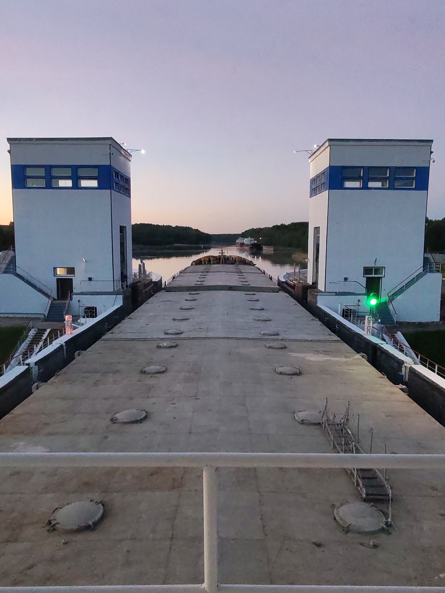 The image depicts a wide view of a navigable waterway lock system at dusk or twilight. Two prominent, identical control towers, painted white with a horizontal blue band and multiple windows, stand symmetrically on either side of a lock chamber. Each tower features exterior lighting fixtures and staircases leading to lower entrances.
In the foreground, the flat, wide deck of a large vessel, possibly a barge, fills the lower half of the frame. This deck is light grey and shows numerous circular hatch covers. Towards the right, a small metal scaffolding or ladder-like structure is visible on the deck. A white railing crosses the very bottom of the image, suggesting the photograph was taken from this vessel or an adjacent platform.
Beyond the lock gates, the waterway extends into the distance, flanked by forested banks. Another, smaller vessel can be faintly seen on the water further down the canal or river.
The sky is transitioning through twilight hues, displaying soft purple overhead blending into warmer orange and pink tones closer to the horizon. Artificial lights are visible, with a prominent green signal light illuminated on the right control tower, indicating permission to proceed. Below this green light, the Russian word "СТОП" (STOP) is visible on a sign. A fainter red or orange light can be seen near the base of the left tower. No people are visible in the scene. The overall atmosphere is calm, highlighting industrial infrastructure for water transportation.