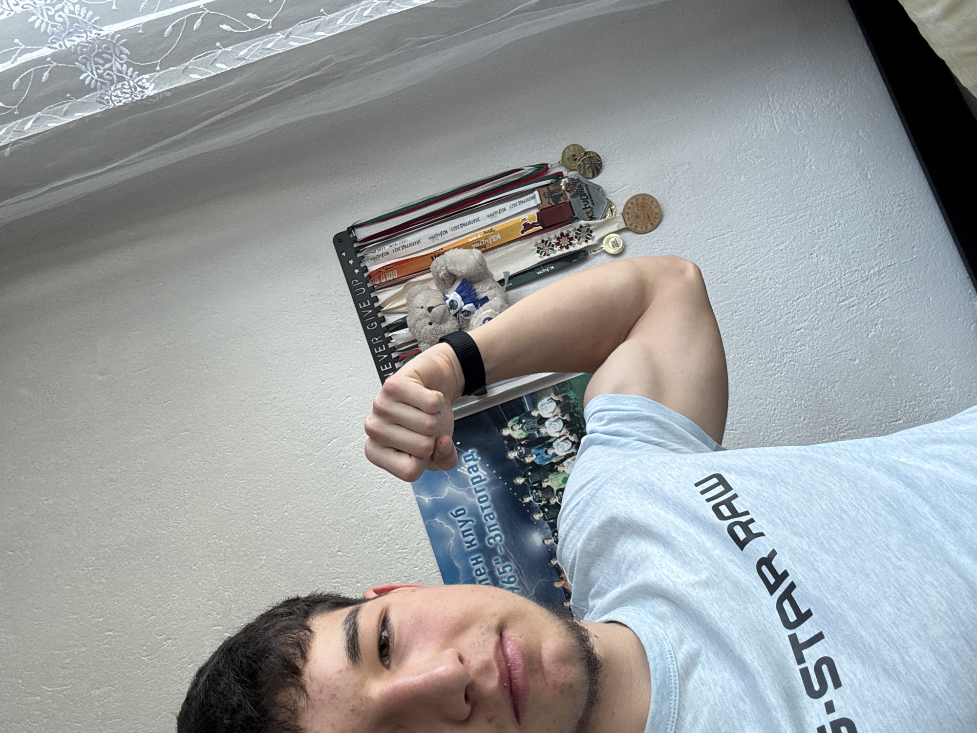 The image is a selfie of a young man wearing a light grey t-shirt with the words "G-STAR RAW" printed on it in black. He is flexing his bicep, which has a black band around the wrist. Behind him is a wall with a medal display. The medals are hung on a black metal rack with the words "NEVER GIVE UP" on it. The display also has two stuffed teddy bears. Below the medal rack, there is a poster of a team, and it's a poster for a team from the "165"-Златоград", as evidenced by the inscription. Златоград (Zlatograd) is a city in Bulgaria. The background includes a partial view of a window with a white curtain, and part of a dark piece of furniture.