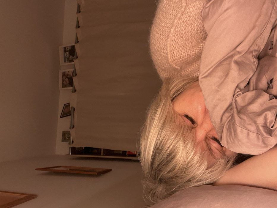 The image shows a person in bed, mostly covered by a pink blanket, with a pink knitted hat on their head. Only their face is visible, and they have white hair. The person's eyes are closed. The room is dimly lit. A curtain hangs on the left side, with small framed pictures attached to it. Two empty wooden picture frames lie on the wall at the bottom left.