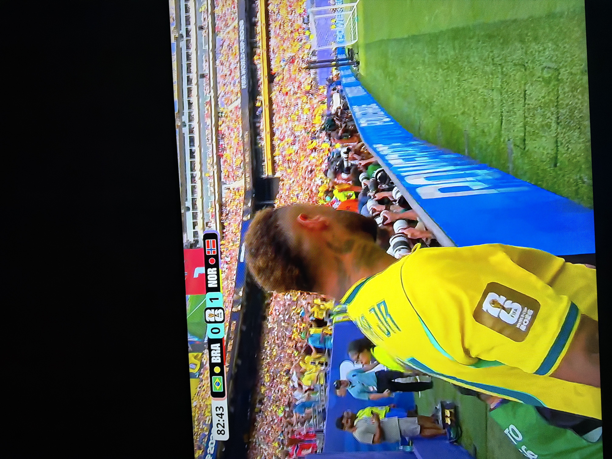The image shows a soccer match with the score displayed as BRA 0 - 1 NOR at 82:43. The game is being played in a stadium with a large crowd in the stands. In the foreground, a player in a yellow jersey with the FIFA World Cup 2026 patch is visible. Several photographers are gathered along the sideline, and some people are standing near a blue barrier. The location is identified as Ognianovo, Bulgaria. The overall scene depicts a live sporting event with players, spectators, media, and stadium infrastructure.