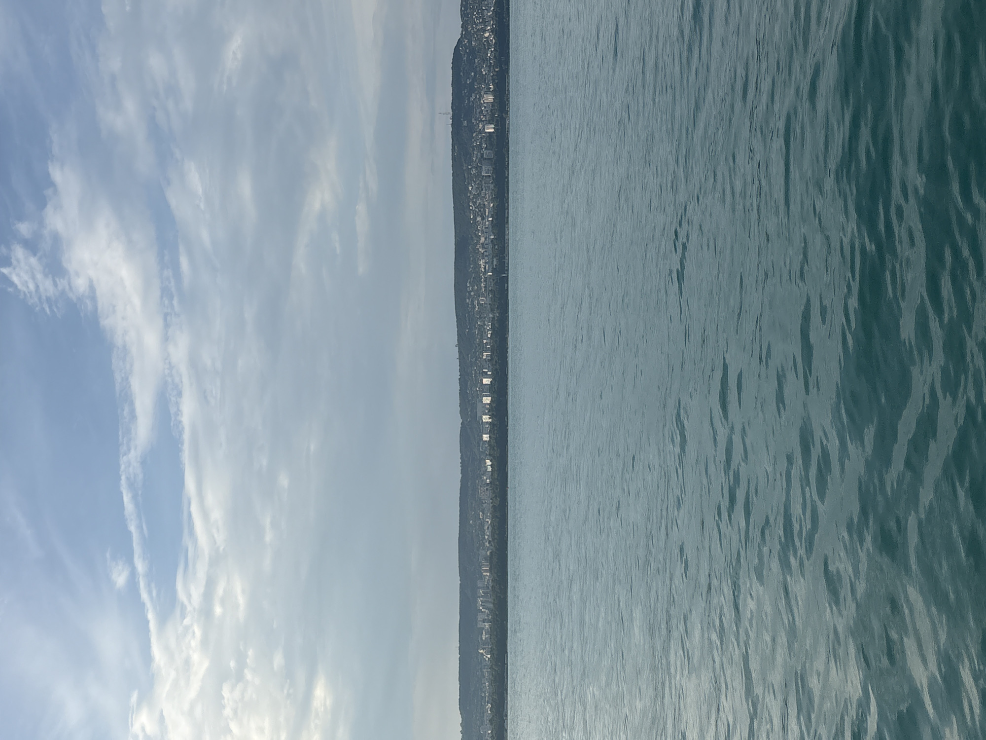 The image presents a wide, horizontal landscape view composed of three distinct layers: sky, water, and a distant city on a landmass. The upper portion of the frame is occupied by a light blue sky with varying formations of white and grey clouds, including both thin, wispy streaks and more diffused, scattered masses. Below the sky, a calm, expansive body of water, colored in shades of turquoise and deep teal, fills the lower two-thirds of the image. The water's surface is gently rippled, creating subtle patterns of light and shadow, with no visible objects or vessels. At the mid-horizontal line, a distant landmass, likely a hill or coastline, stretches across the entire width of the frame, separating the sky and water. This landmass is densely covered with dark green vegetation, forming a tree line immediately bordering the water. Above this vegetation, a densely built-up urban area is visible, with numerous buildings of various sizes extending up the slope of the landmass. Several structures, particularly on the right side of the city, appear to have bright, reflective facades. A tall, slender antenna or communication tower is discernible on the highest point of the landmass, towards the right. There are no individuals or discernible human activity depicted in the foreground or on the water. The scene is illuminated by natural daylight. The location is an unknown city near a large body of water.