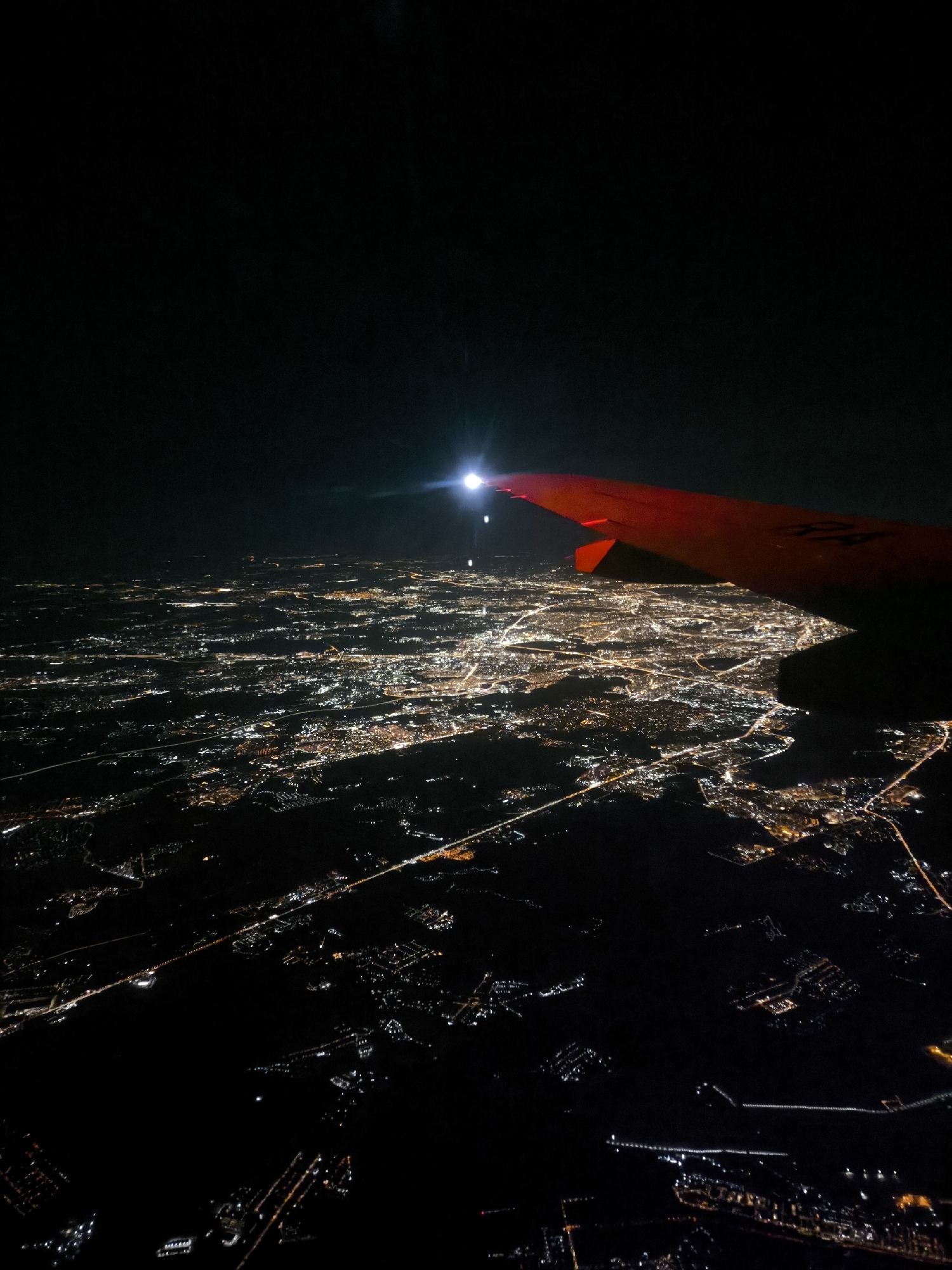 An aerial view from an airplane at night shows the cityscape of Khimki, Russia. The city lights create a web of yellow and white illumination across the dark landscape. Part of the airplane wing, colored red, is visible in the upper right of the frame, with a bright light emanating from its edge. The night sky is dark and clear.