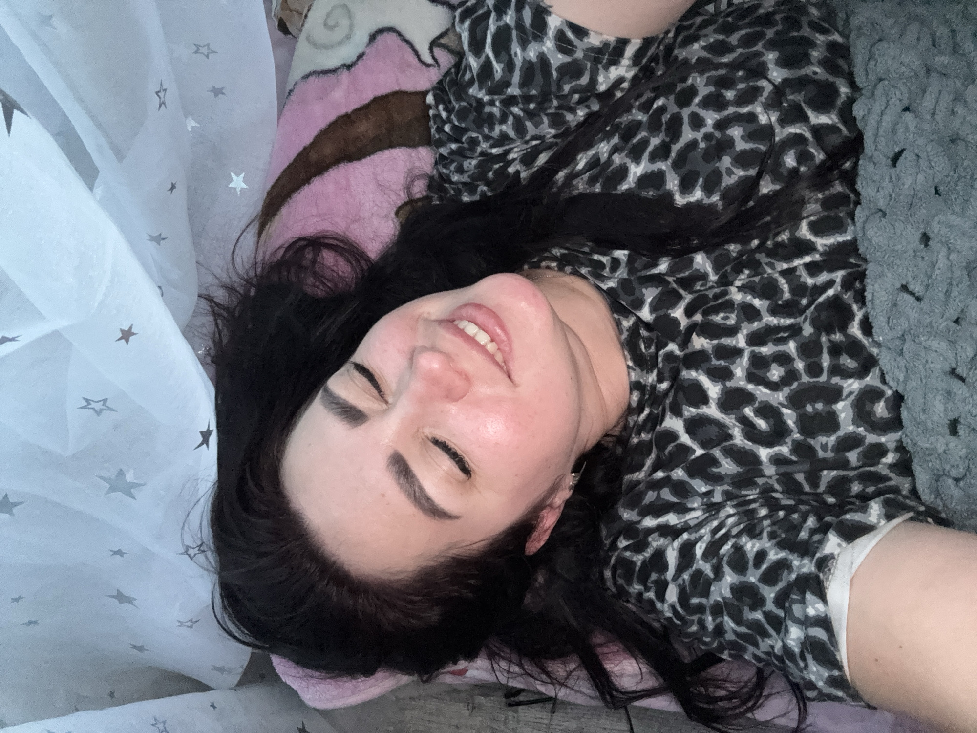 A woman with long, dark hair is lying down on a bed with a star-patterned sheet and a pink blanket. She is wearing a leopard-print top and has her eyes closed, with a smile on her face. A gray, chunky knit blanket is draped over her body. The scene appears to be indoors.