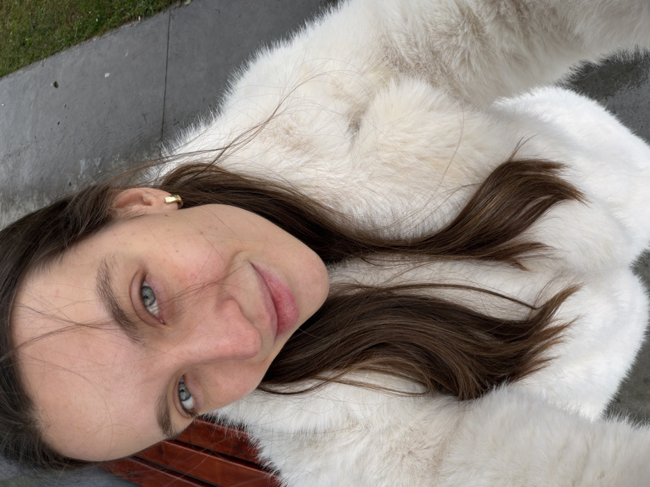 A fair-skinned woman with brown hair and blue eyes is captured in a close-up shot, tilted to the side. She's wearing a white fur coat, with portions of the coat visible beside her. A gold earring is visible in her left ear. The background shows a glimpse of green grass and gray concrete, potentially a sidewalk or walkway. The subject's expression is relaxed and inviting.