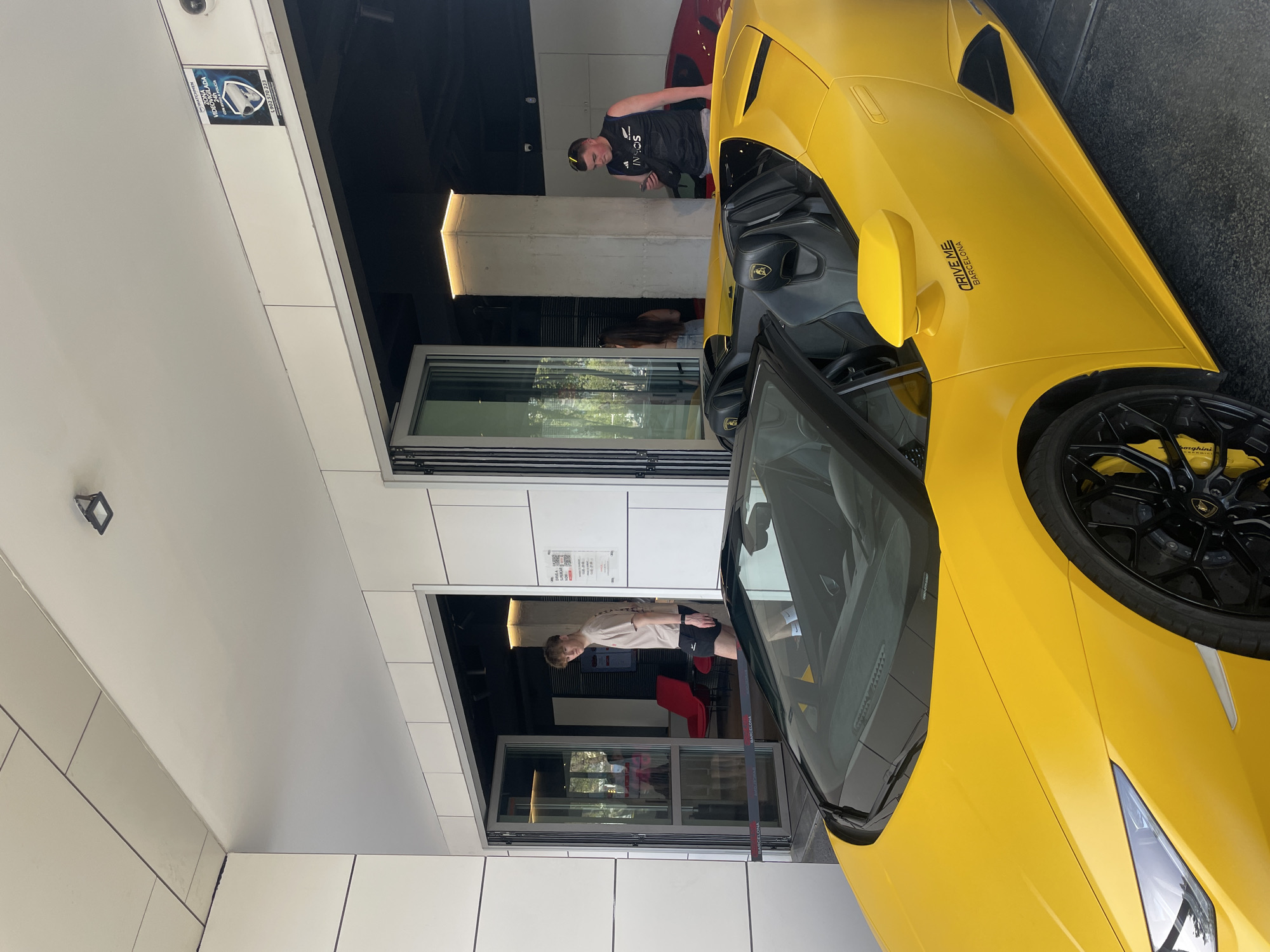 A medium, eye-level shot shows the front of a yellow Lamborghini in Barcelona, Spain, parked in front of a modern building with white, rectangular tiling. The car's driver's side door is open, revealing a black leather interior. A sticker on the car reads "DRIVE ME Barcelona." The black wheels feature yellow brake calipers with the Lamborghini logo.
Two men are visible in the background, standing in the building's open doorway. One man, wearing a black t-shirt and baseball cap, appears to be looking at his cell phone. The other man, wearing a light-colored t-shirt and black shorts, stands near a red chair. The building has two tiers of sliding glass doors. A small security camera and a sign are affixed to the tiled wall above the doorway.