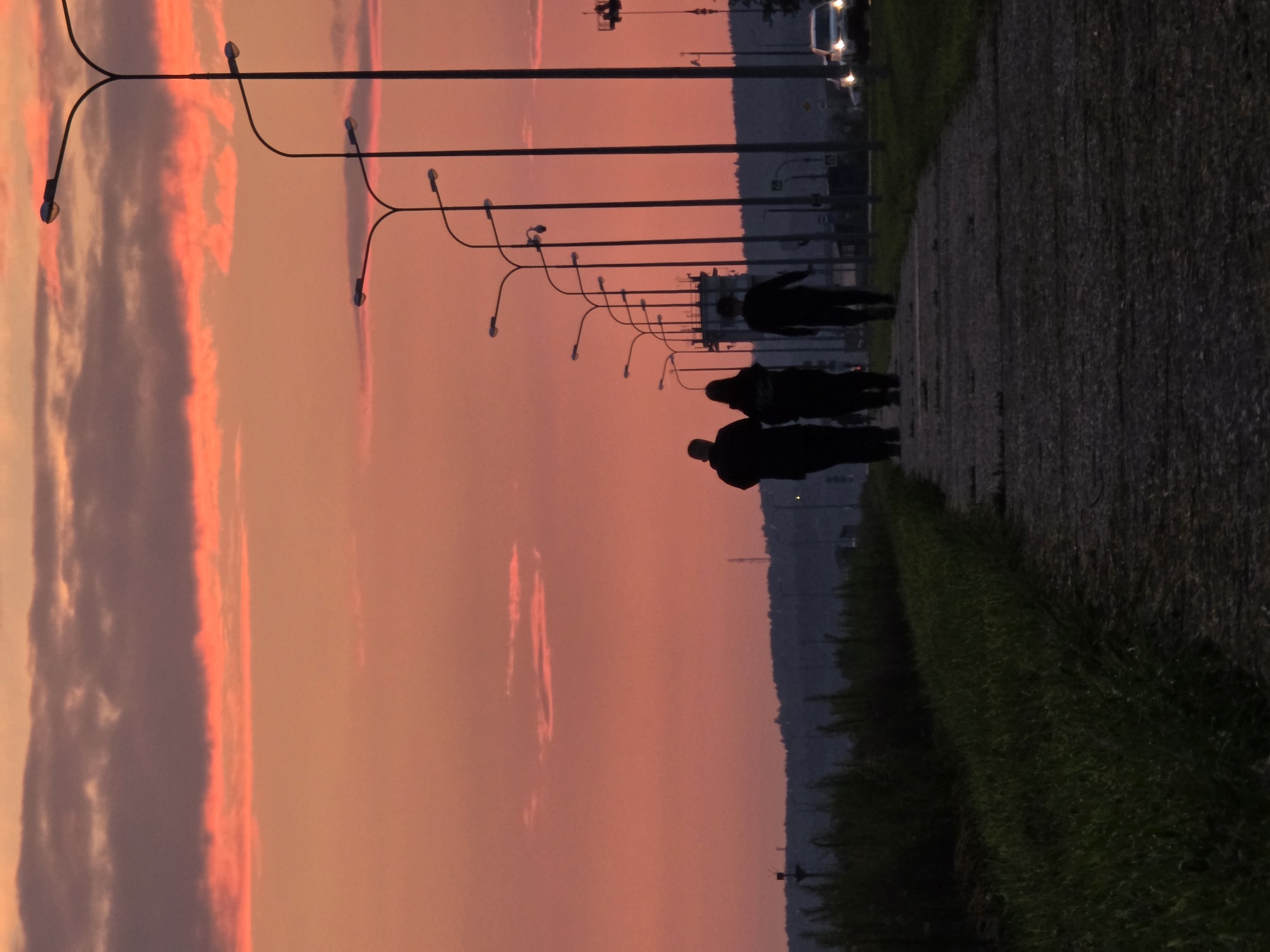 The image depicts a pedestrian walkway alongside a road, bordered by grass on the left and lamp posts on the right. Three individuals, silhouetted against a vibrant sunset, are walking away from the camera on the pathway. The sky is a dramatic gradient of orange and pink, with dark clouds scattered across it. In the background, a line of trees and distant structures, possibly industrial buildings or housing, are visible. A white car with its headlights on is approaching on the road. The scene appears to be located on the outskirts of Chaikovsky, Russia, given the context. The overall atmosphere is serene, with the setting sun casting long shadows and creating a warm, diffused light.