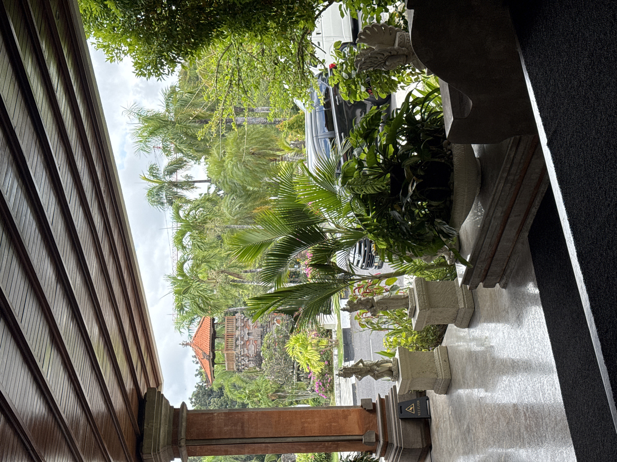 The image depicts an outdoor scene from a covered walkway or entrance area, likely in a tropical or sub-tropical location, possibly Kuta Selatan, Indonesia, given the architectural style and vegetation. The foreground features a polished, reflective floor with a dark mat leading down. To the left, a thick, reddish-brown pillar supports a wooden slatted ceiling. Two stone statues of figures in traditional dress stand on pedestals on either side of a pathway. A "WET FLOOR" sign is visible near the pillar. Beyond the covered area, lush green palm trees and other tropical foliage dominate the landscape. In the middle ground, a multi-tiered stone structure with an orange-tiled roof, reminiscent of a Balinese temple or pavilion, is visible. A black SUV and a white van are parked on a road that runs behind the statues and garden. The sky is overcast. The overall impression is one of an upscale resort or a culturally significant site with tropical landscaping.