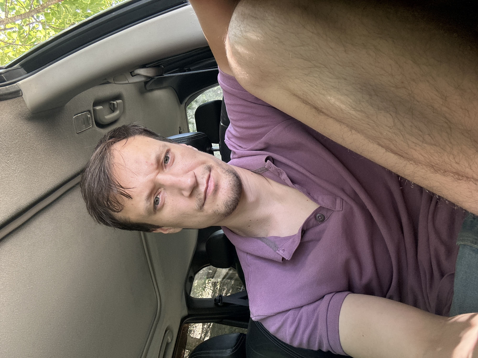 A man sits in the driver's seat of a car, looking at the camera. He has short, brown hair and is wearing a purple polo shirt. The car's interior is visible, including the headliner, dashboard, and seats. Sunlight streams in through the windows, illuminating the man's legs and the car's interior. The man's left arm is resting on his leg. The scene appears to be outdoors, as evidenced by the trees and foliage visible through the car windows. It is daytime, and the weather appears to be warm. The location is identified as Makiv, Ukraine.