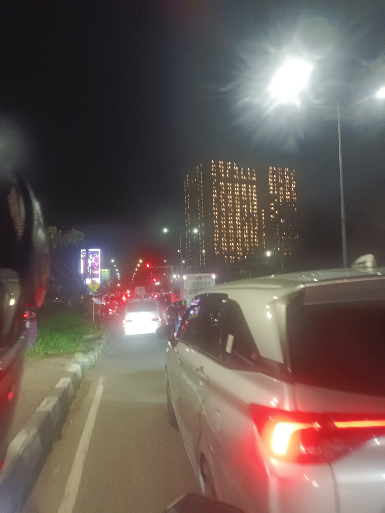 The image depicts a night scene in an urban environment, specifically a traffic jam. The viewpoint is likely from within a vehicle, as part of a helmet and the rear of another vehicle are visible in the foreground.
Several cars, trucks, and motorcycles are congested on the road, their headlights and taillights illuminating the area. The traffic appears to be at a standstill or moving slowly.
On the left side of the road, there's a strip of greenery and a sidewalk. A large billboard is also visible.
In the background, a tall building is illuminated with a pattern of lights, potentially forming a message or an image. Streetlights are also present, contributing to the overall illumination of the scene.