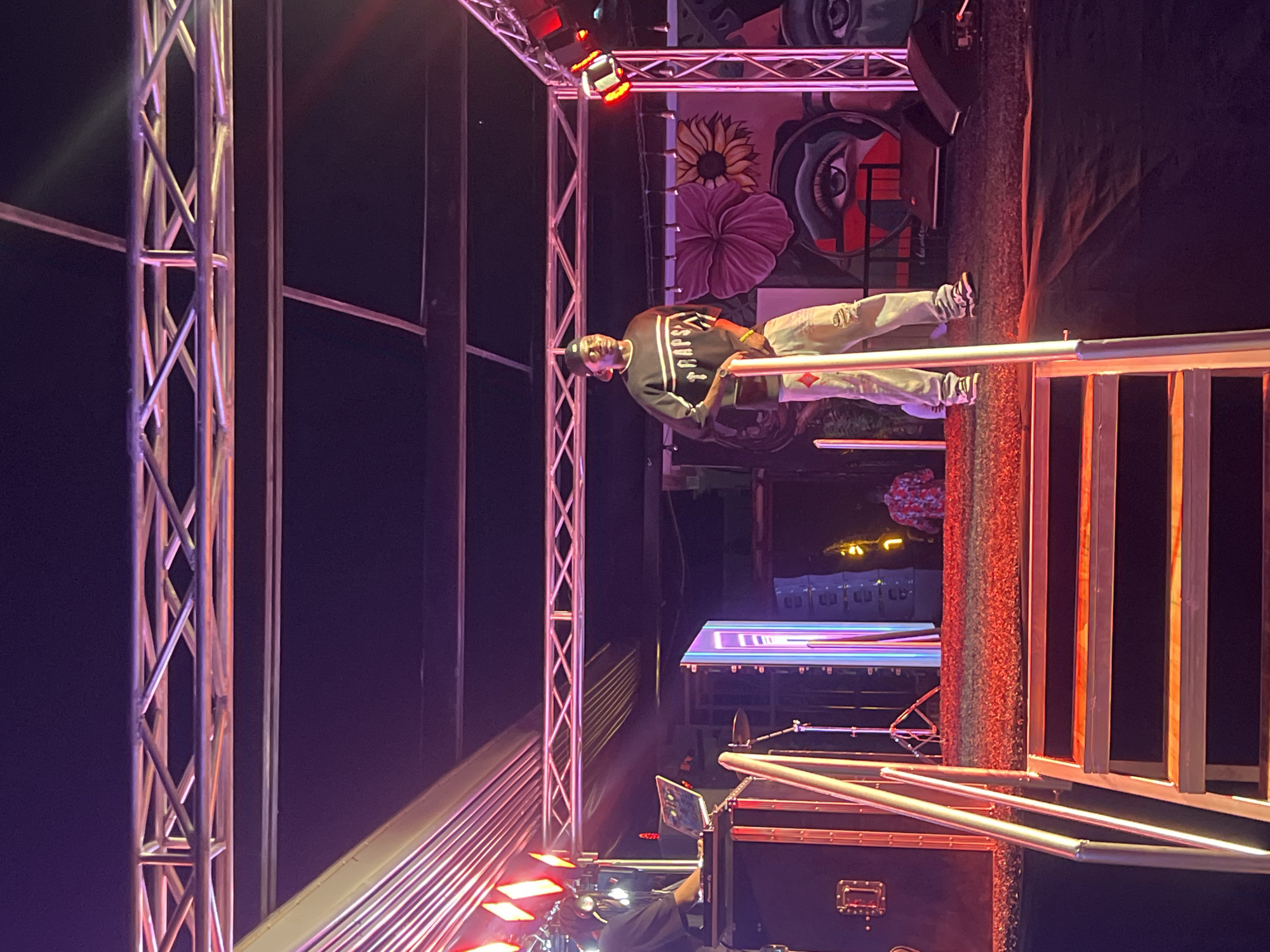 The image depicts a scene at night in Howayi, Nigeria. A man wearing a black "Traps" sweater, light-colored pants, and a baseball cap is standing on a structure made of metal bars. Behind him is a backdrop with artwork including flowers and an eye. To the left, there are more metal structural supports. Below and in front of him are stage elements including what appears to be a mixing console and metal railings. A building is visible in the background. The overall lighting is a mixture of red, pink, and purple.