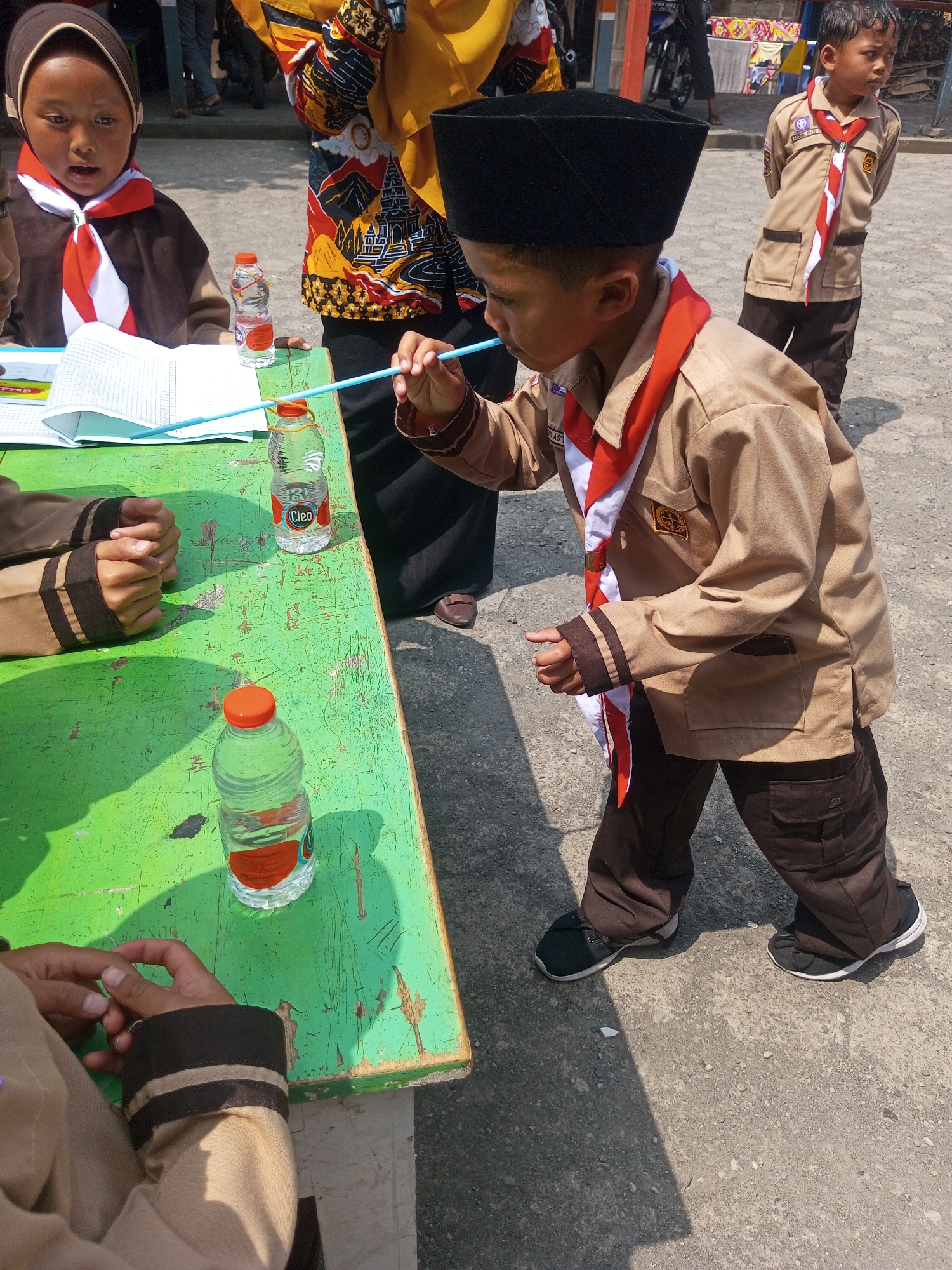 In the city of Kalibebe, Indonesia, on a bright, sunny day, a young boy wearing a black cap and a khaki scout uniform with a red and white sash is captured in the foreground. He is actively drinking from a clear plastic water bottle using a light blue straw. To his left, a green wooden table is present, holding another identical water bottle and an open notebook. On the table, a girl, also in a khaki uniform and headscarf, has her hands resting on the notebook. Behind the boy, another young boy in a similar scout uniform stands, looking away from the camera. The ground is a mix of asphalt and gravel, with shadows indicating the harsh midday sun. The overall scene suggests an outdoor educational or recreational activity.
