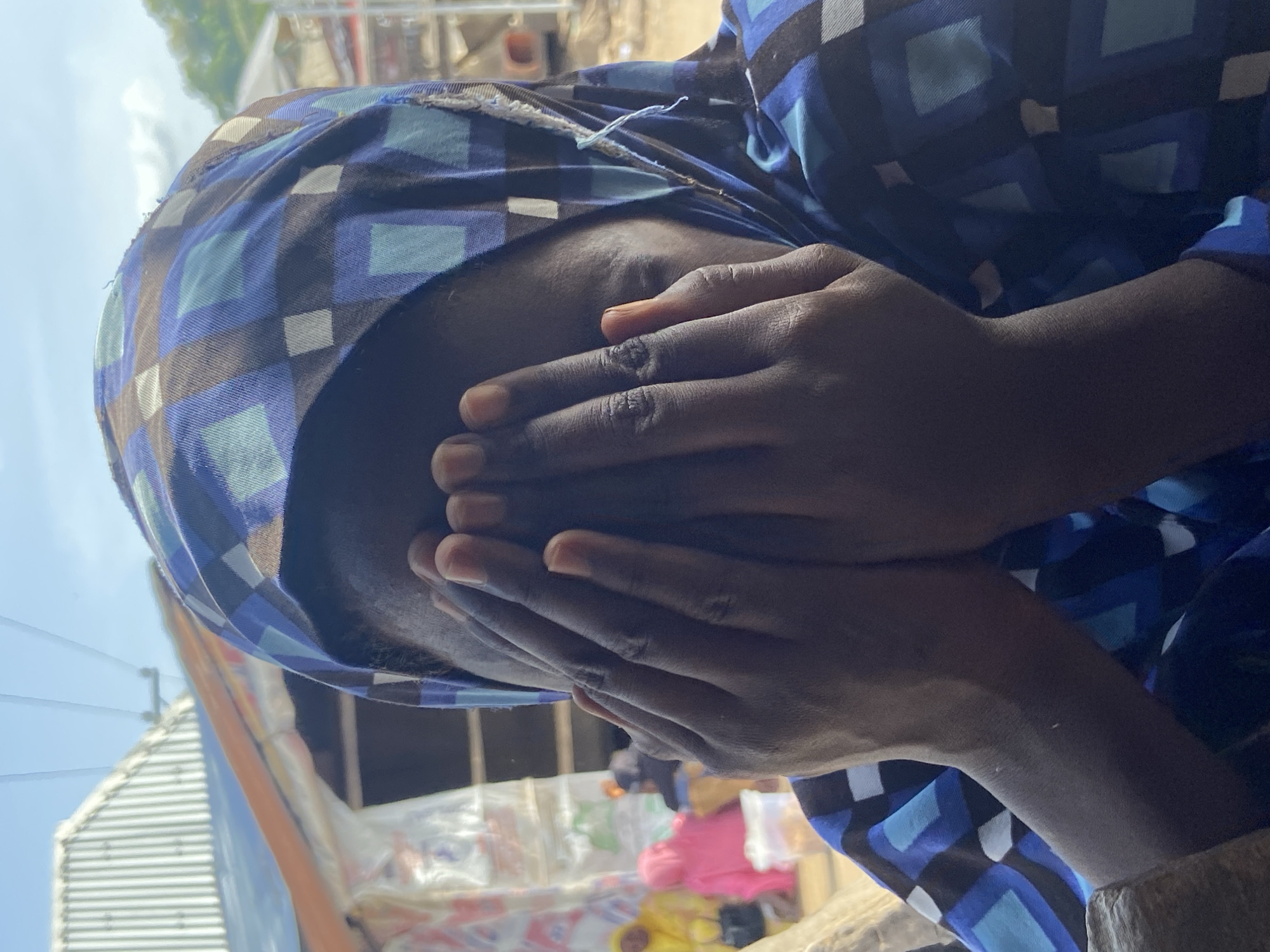 A person wearing a blue patterned headscarf and shirt is covering their face with their hands. The individual's skin tone is dark. The person appears to be inside a partially enclosed space, possibly a shop or market stall, as indicated by visible merchandise in the background, including items wrapped in clear plastic. The scene is located in Mararaba, Nigeria as indicated in the instructions. The lighting is bright, suggesting daytime.