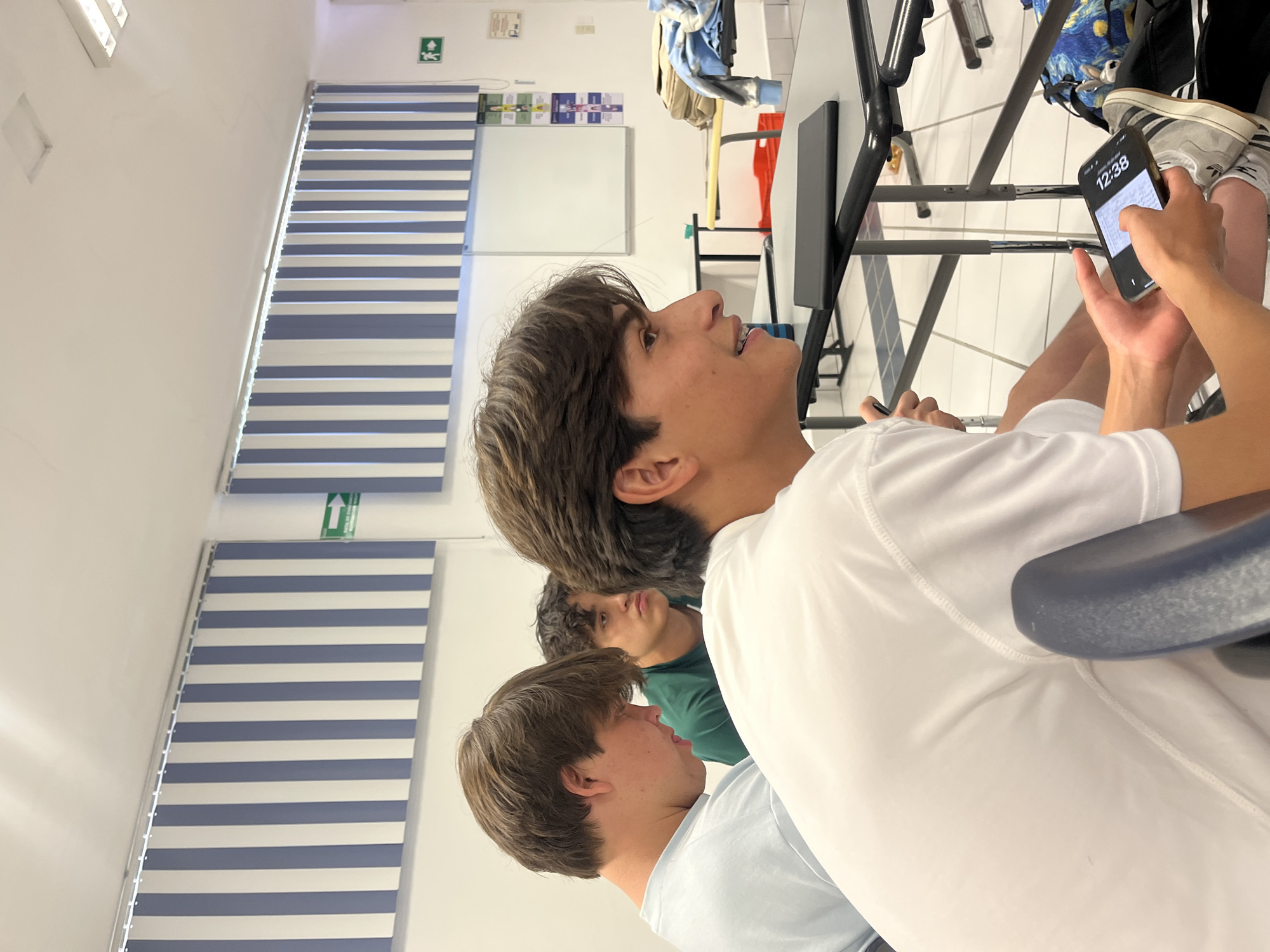 The image depicts an indoor scene, likely a classroom or study hall, featuring three young males. The perspective is from behind and to the side of the closest subject.
In the foreground, a young male with dark brown hair, wearing a white t-shirt, is seated. His head is turned to the right, and he appears to be looking upwards or forward with a slightly open mouth, as if listening or observing. His right hand holds a black smartphone, which clearly displays the time "12:38" on its screen. Below his hand, a backpack with a colorful, swirling pattern resembling Van Gogh's "Starry Night" is visible on the floor or a low shelf.
Behind this individual, another young male with curly dark hair, wearing a dark green t-shirt, is partially visible, also looking in a similar forward direction. Further to the left and slightly below, a third young male in a light blue t-shirt is visible, with only part of his head and shoulder shown.
The setting includes white walls and a white ceiling with fluorescent light fixtures. On the left side of the image, two windows are covered with horizontal blue and white striped blinds. A green emergency exit sign with a white arrow pointing upwards is mounted on the wall above the blinds. Further into the background, a whiteboard with some papers attached to it is visible, and black metal desks or tables are arranged.
The overall impression is of a quiet, possibly instructional or study-focused environment during midday.