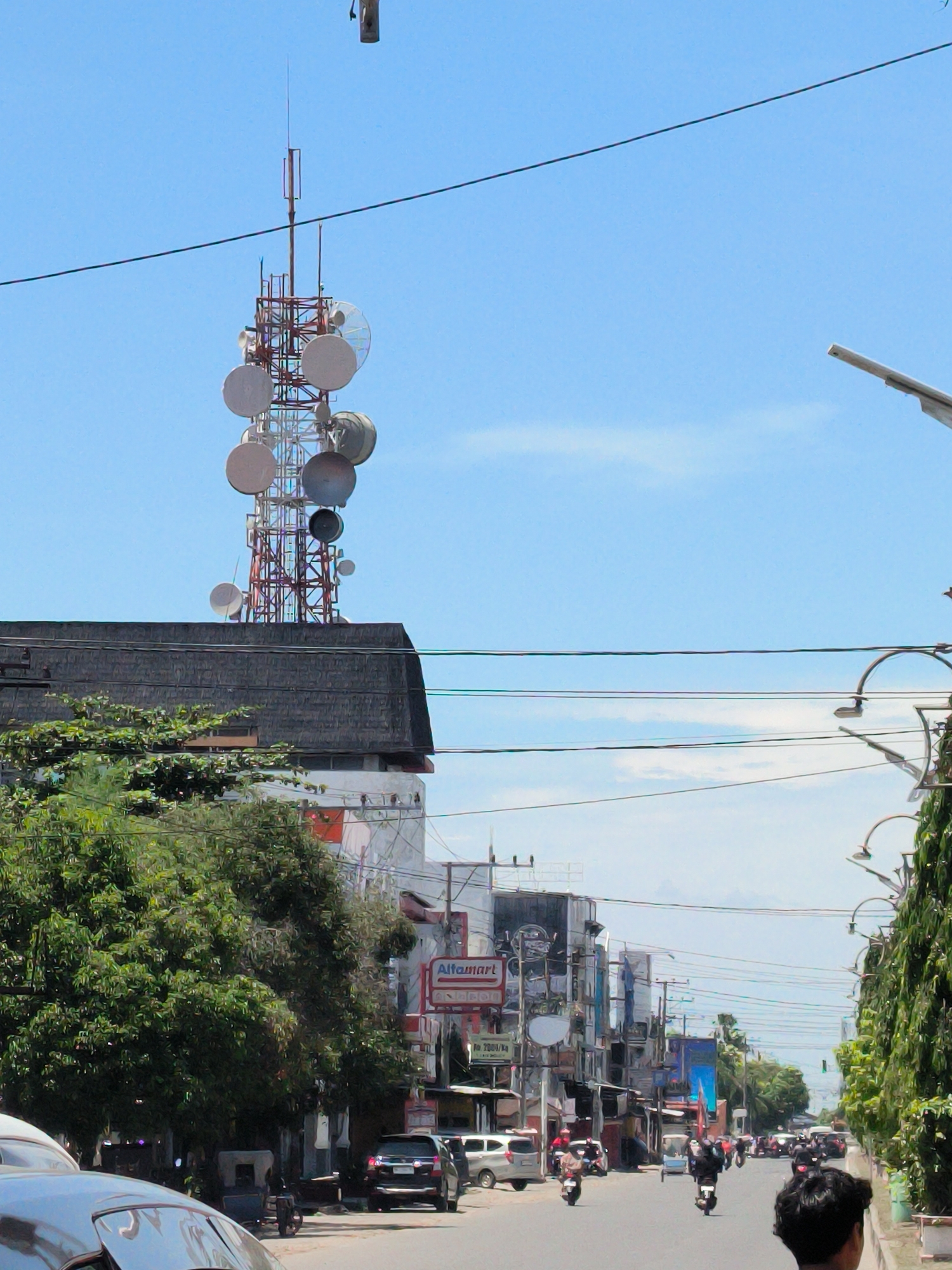 The image depicts a street scene in Lhokseumawe, Indonesia, during daylight. A prominent telecommunications tower with multiple satellite dishes sits atop a building. The street is lined with buildings, including a visible "Alfamart" store. Cars and motorcycles populate the street. There are trees on either side of the road and overhead power lines. Several people are walking along the street. The sky is blue with sparse clouds.