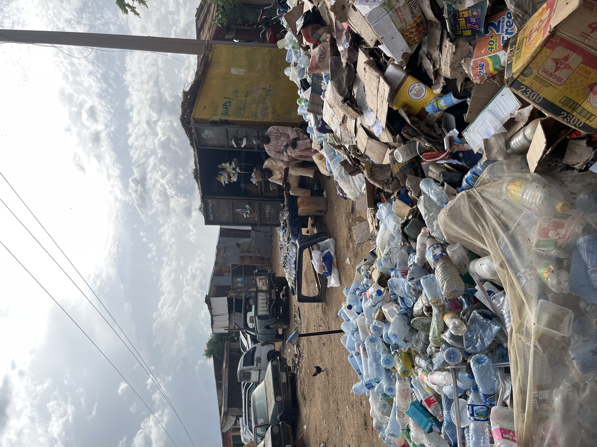 In Jos, Nigeria, a scene unfolds at a roadside recycling yard, characterized by a large pile of discarded plastic bottles and cardboard boxes dominating the foreground. The ground is dusty and uneven, indicative of an outdoor, unpaved area. In the mid-ground, several parked vehicles, including cars and a truck, suggest a bustling commercial or transportation hub. A small chicken pecks at the ground near a weighing scale, adding a touch of rural life to the urban setting. Behind the pile of recyclables, a yellow shipping container serves as a makeshift shop, its open door revealing shelves stocked with footwear. Two men, dressed in traditional attire, sit in front of the container, possibly vendors or shopkeepers. The background reveals more corrugated metal structures, characteristic of informal settlements or market stalls. The sky is partly cloudy, with visible power lines and a utility pole stretching upwards, indicating the presence of infrastructure. The overall impression is one of resourcefulness and informal economy operating amidst a backdrop of urban development and environmental challenges.