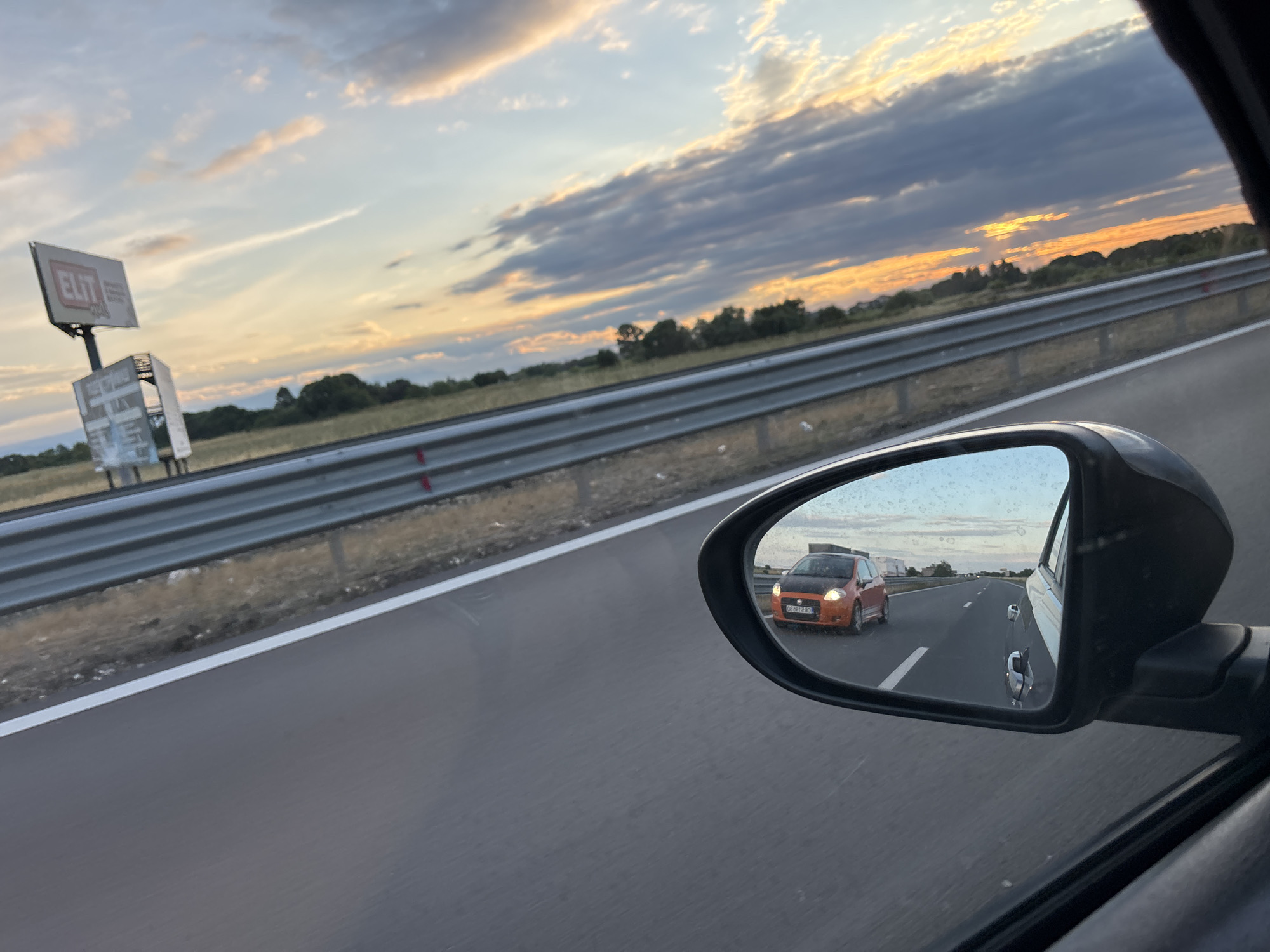 A side-view mirror of a vehicle reflects an orange hatchback car with a black hood driving on a highway. The car in the mirror has a license plate that reads "08 BB 2183". The highway is bordered by a metal guardrail and dry grass. In the distance, trees line the horizon under a sky with dramatic clouds illuminated by the setting sun. To the left of the highway, a billboard with the word "ELIT" is visible. The image was taken on the highway near Kostievo, Bulgaria.
