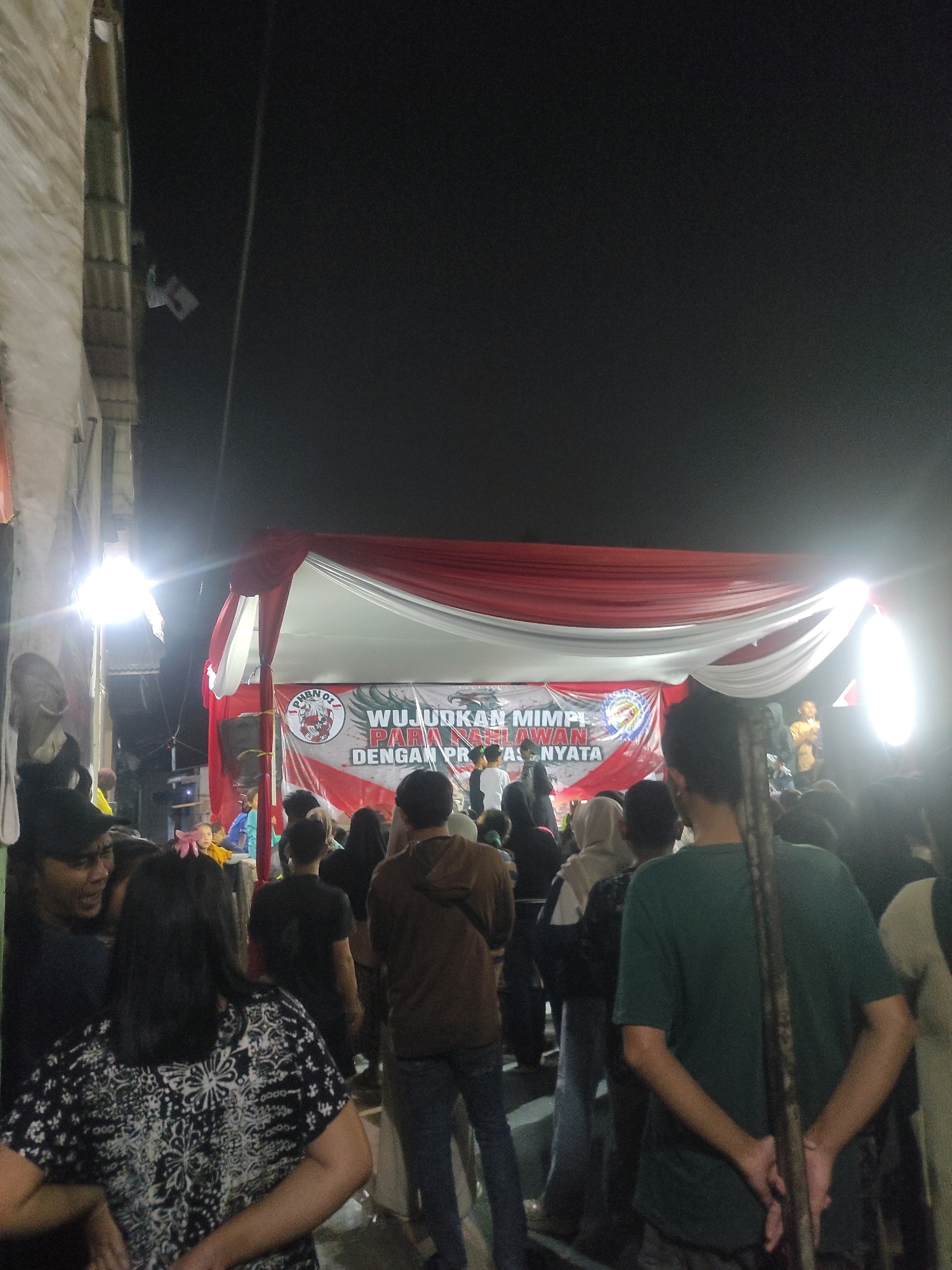 In Padalarang, Indonesia, a nighttime outdoor event is taking place under a large red and white tent. A crowd of people, seen from behind and partially from the side, are gathered. In the background, a banner is displayed with Indonesian text and a circular logo containing the letters "PMDN 01." The lighting appears to be artificial, with bright lights illuminating the tent area and creating glare. The overall atmosphere suggests a community gathering or performance.