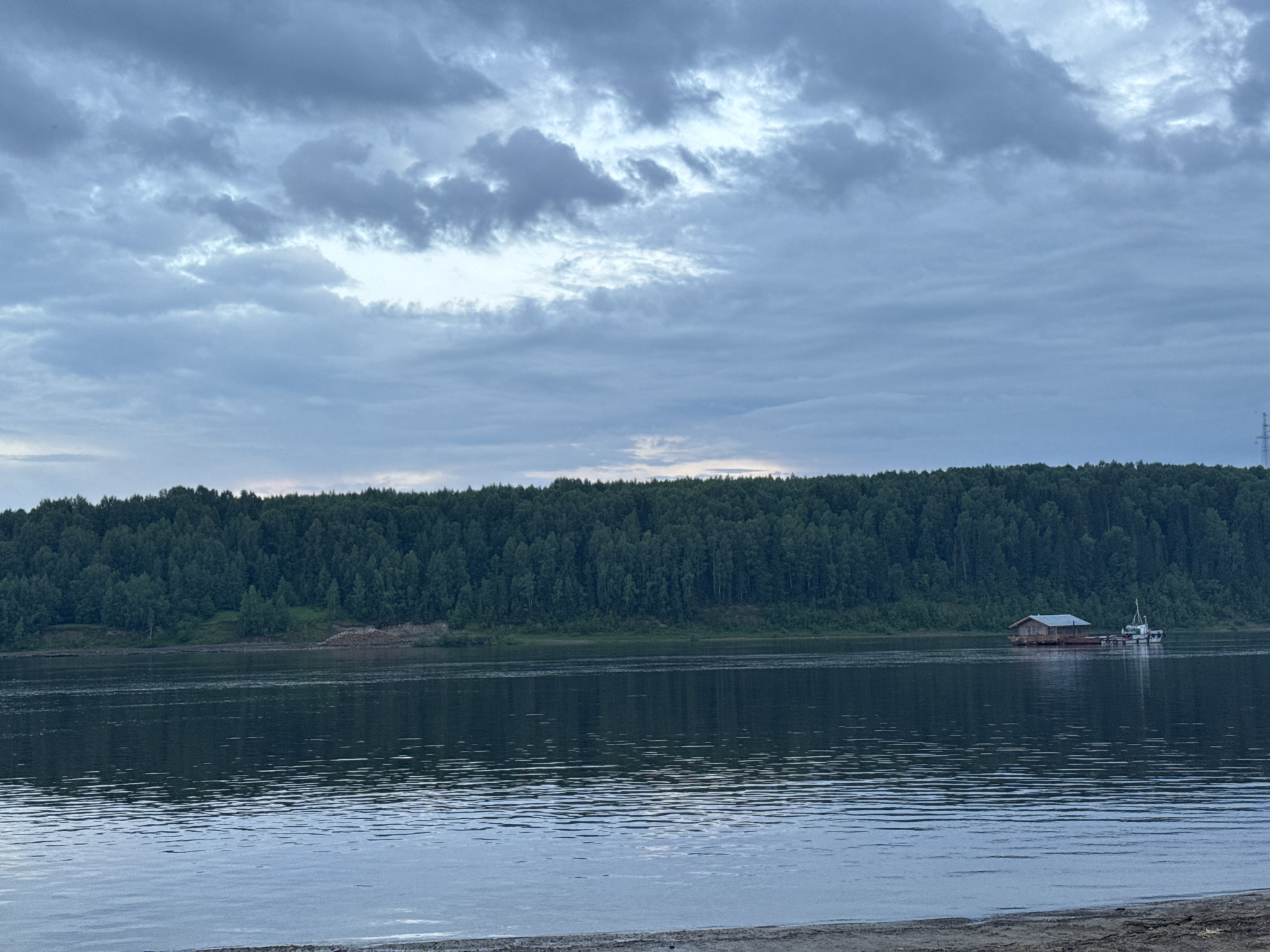 A wide, dark river flows toward the viewer, reflecting a cloudy, pale blue sky and a dense, dark green forest that lines the opposite bank. In the middle of the river, a barge with a small wooden structure on it, possibly a dwelling or storage shed, is moored. A small white boat is attached to the rear of the barge. The barge is positioned to the right of the center of the frame, with the forest extending across the background. The foreground shows a sandy bank. The image appears to be taken in or near Tyulkino, Russia.