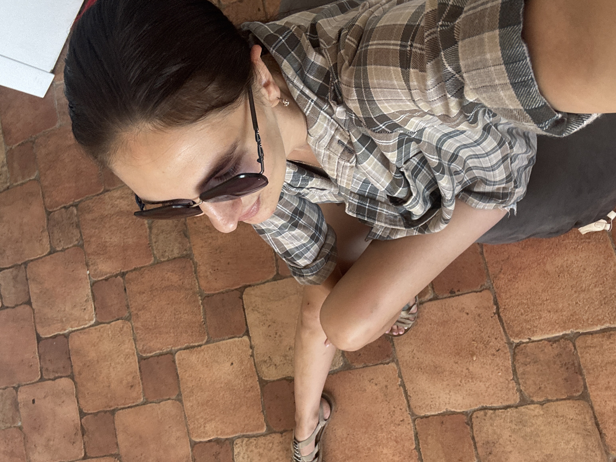 A woman with dark hair is sitting on a floor made of terracotta tiles. She is wearing sunglasses and a plaid shirt. Her left leg is extended, and her right leg is bent. She is wearing sandals on both feet. The lighting suggests it is daytime, and the setting appears to be indoors or a covered outdoor area.
