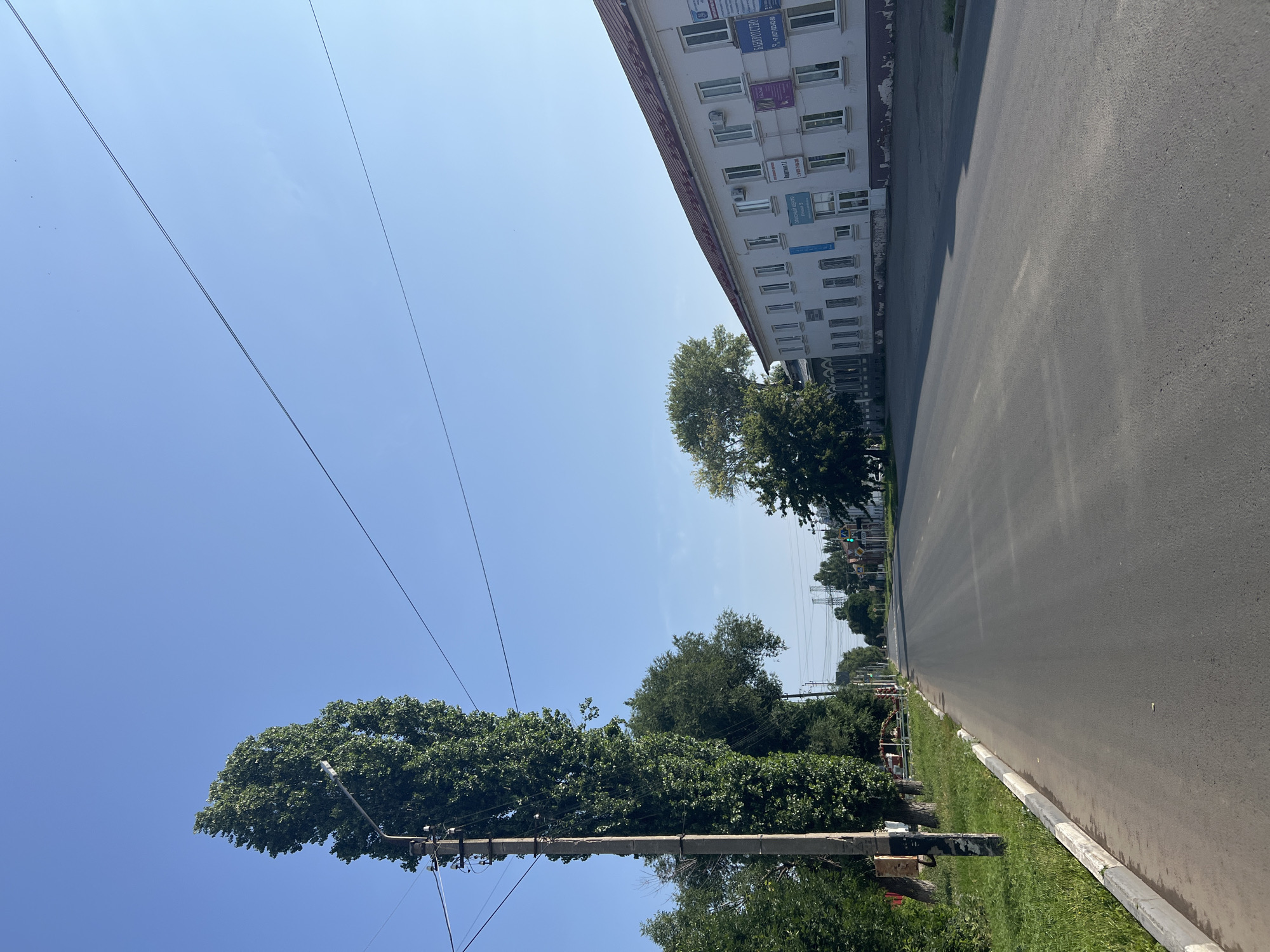 The image depicts a view of a street in the city of Balakovo, Russia, on a clear, sunny day. The foreground shows a paved road with a slight sheen, suggesting it may be newly paved or recently rained on. A grey curb with white and black stripes runs along the left side of the road, bordered by a strip of green grass. To the left, a tall, slender poplar tree stands next to a concrete utility pole adorned with wires and what appear to be electrical components. Behind the utility pole and tree, more trees are visible, creating a dense green line.
In the mid-ground, the road continues straight into the distance, flanked by more trees and buildings. On the right side of the road, a long, two-story building with a red roof is prominent. The building is painted white and features numerous windows and air conditioning units. Several signs are affixed to the building's facade, including one that appears to be for a "Bankrotstvo" (Bankruptcy) service, and another with Cyrillic text. The building has a slightly weathered appearance, with some peeling paint visible at the base.
Further down the street, power lines are suspended overhead, and in the far distance, large industrial towers can be seen against the clear blue sky. The overall impression is of a typical urban or semi-urban street scene in Russia, characterized by utilitarian architecture, abundant trees, and the presence of infrastructure. There are no people visible in the image, and no specific activity is taking place. The lighting suggests it is likely daytime, with the sun casting subtle shadows.