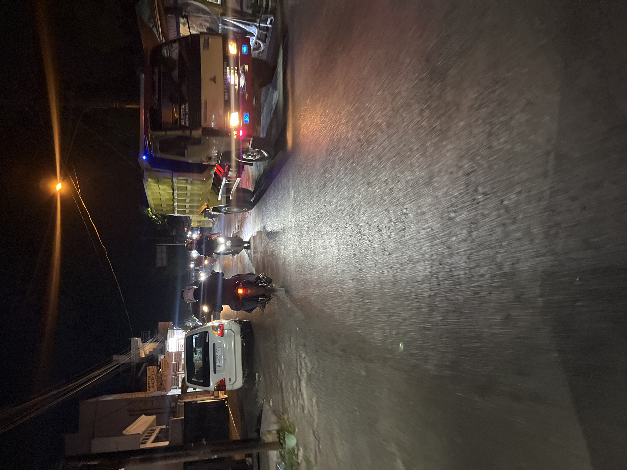 At night on a wet asphalt road in Pecangaan, Indonesia, a white minivan with license plate "D 1322 GG" is parked on the left side. A person rides a motorcycle behind the minivan, heading away from the camera. To the right of the minivan, a yellow dump truck with blue flashing lights is moving forward, its headlights illuminated. Further down the road, another motorcycle is visible, also moving forward. The scene is illuminated by streetlights and vehicle lights. The surrounding area features buildings and utility poles. The road surface is reflective due to the wet conditions, creating streaks of light.