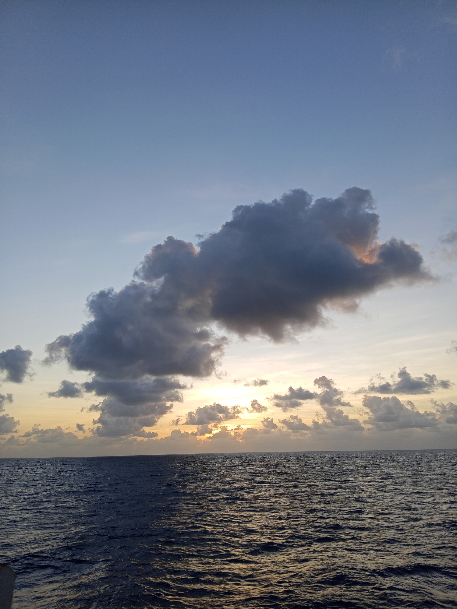 The image depicts a wide expanse of dark blue ocean under a pale blue sky during what appears to be either sunrise or sunset. A line of clouds stretches across the horizon, with the sun peeking through in the distance, casting a warm glow. A large, dark cloud formation dominates the upper middle section of the sky. The water is slightly rippled, reflecting the subdued light. There are no people or man-made structures visible in the frame, suggesting an open-sea environment. The scene is tranquil and natural.