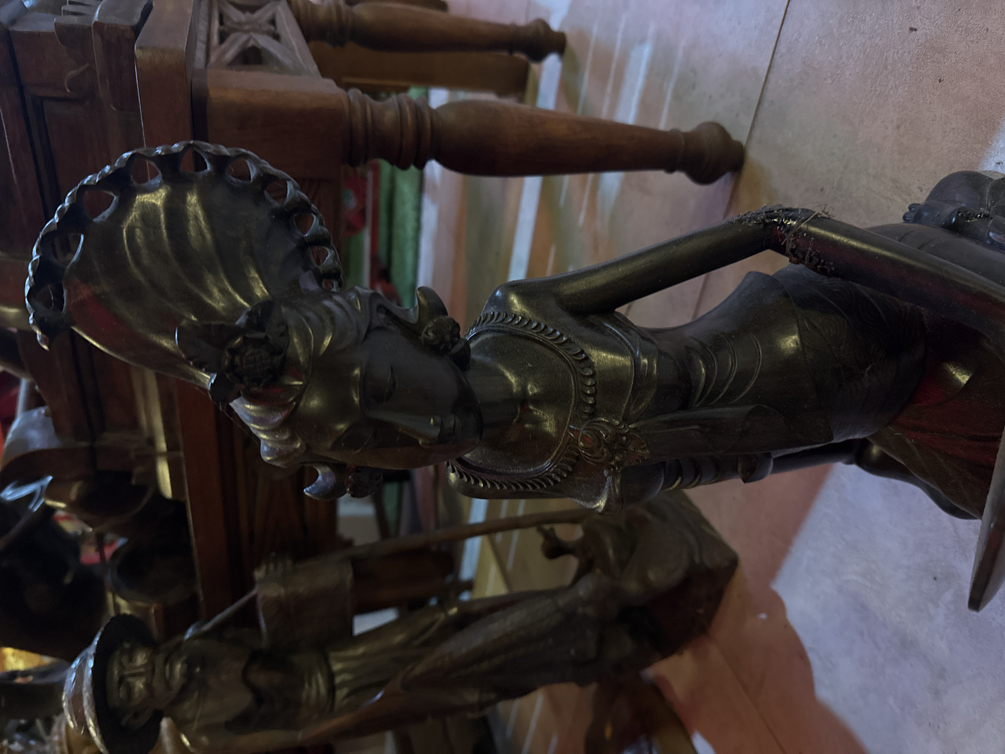 The image shows a dark, possibly bronze or wooden, statue of a reclining figure. The figure is depicted as a person with elaborate headwear that fans out like a peacock's tail, adorned with carved details and holes. The statue is intricately crafted, featuring delicate jewelry around the neck and a detailed garment. The figure's body is slender and posed in a relaxed, reclining position.
The statue is placed on a wooden surface, likely furniture, with visible carved details. In the background, parts of more wooden structures and what appear to be other statues or decorative elements can be seen. The lighting suggests an indoor setting, possibly a museum, gallery, or a home with ornate decor. The colors are muted, dominated by dark tones of the statue and the rich browns of the wood. The overall impression is one of artistry and perhaps cultural significance, given the elaborate ornamentation. The image is oriented vertically, with the statue appearing to recline from left to right. The Kuta, Indonesia location context suggests the statue might be of Balinese or Indonesian origin, possibly representing a deity, mythical figure, or a depiction of traditional life.