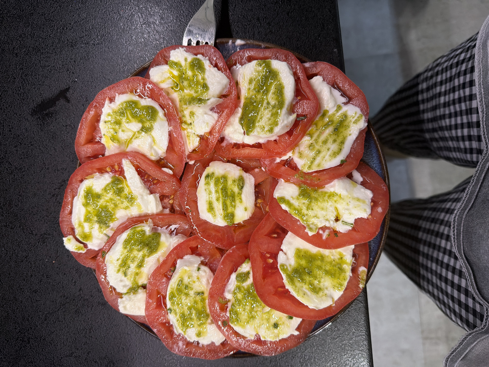 A top-down shot shows a plate of Caprese salad consisting of sliced tomatoes and mozzarella drizzled with green pesto, with a fork resting near the edge of the plate. The plate is on a dark surface, possibly a table, and part of a person wearing checkered pants is visible in the background. The location appears to be indoors.