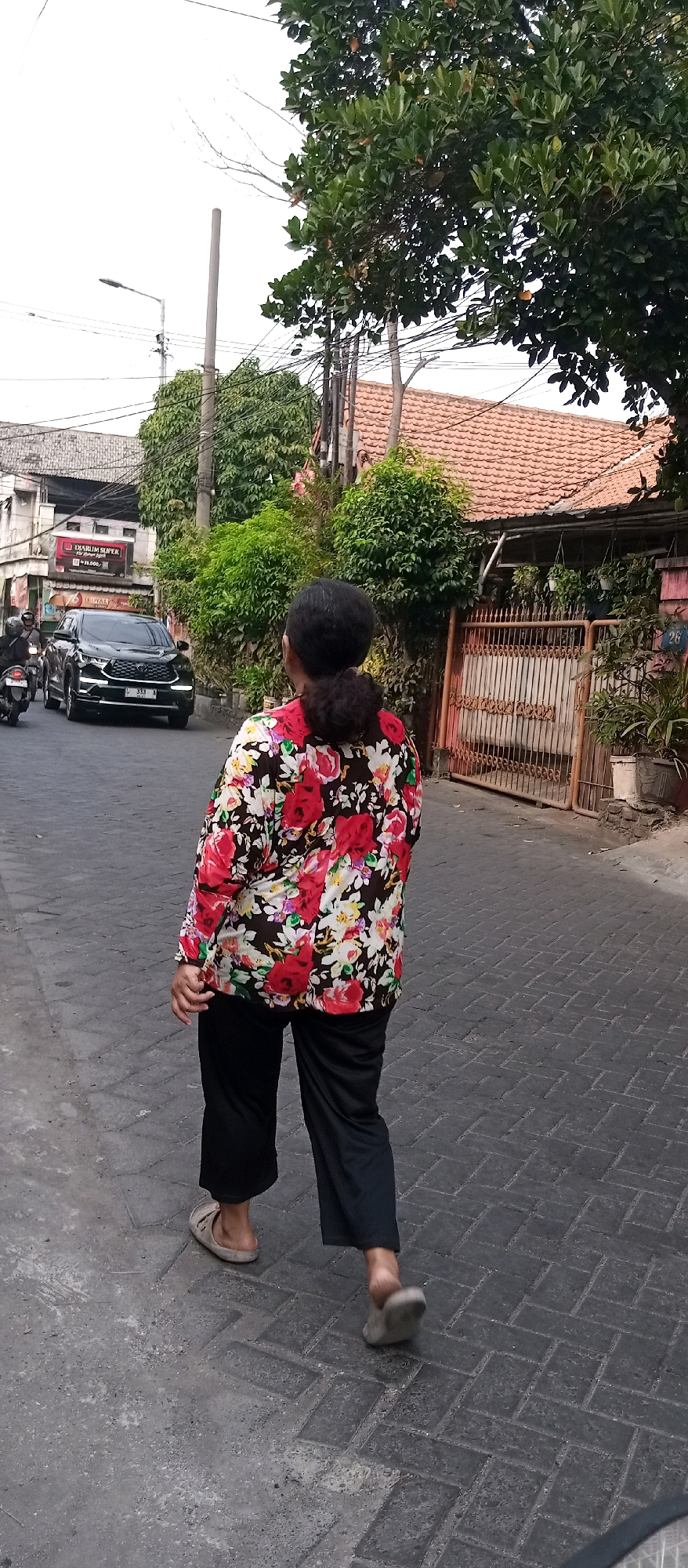 The image depicts a street scene in Wedoro, Indonesia. A woman with dark hair tied in a bun is walking away from the camera on a paved street. She is wearing a floral print blouse with red, white, and pink flowers on a black background, and loose-fitting black pants. Her footwear appears to be simple sandals. To the left of the woman, a black SUV is driving down the street, and a motorcycle is visible behind it. On the right side of the street, there is a property with a red-tiled roof and a metal gate, with plants visible in pots. The street is lined with buildings and lush green trees. The sky is overcast.