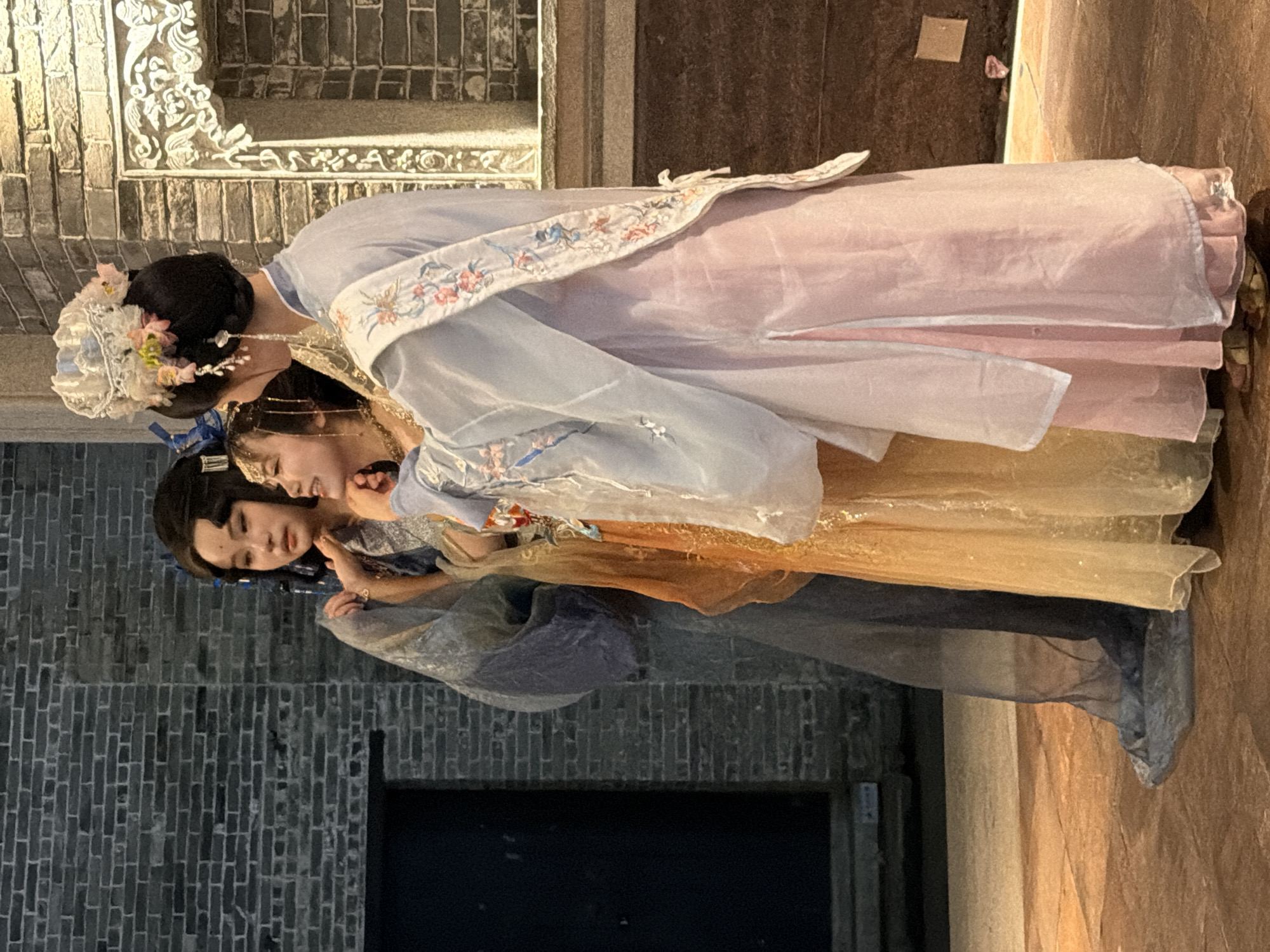 Three individuals in traditional Chinese attire stand closely together in front of a brick wall. The person on the left wears a long, flowing blue garment. The person in the middle is adorned in an orange and gold dress, with elaborate jewelry. The person on the right is dressed in a light blue outer robe over a pink dress, and wears an ornate headdress. They are positioned in an outdoor setting, likely on a stone-paved ground, with a decorative architectural element visible on the right. The overall scene suggests a cultural performance or reenactment. The background consists of a dark brick wall and a large dark doorway or opening on the left. The lighting appears to be artificial, casting warm tones on the ground and figures. The location is identified as Chancheng District, China.