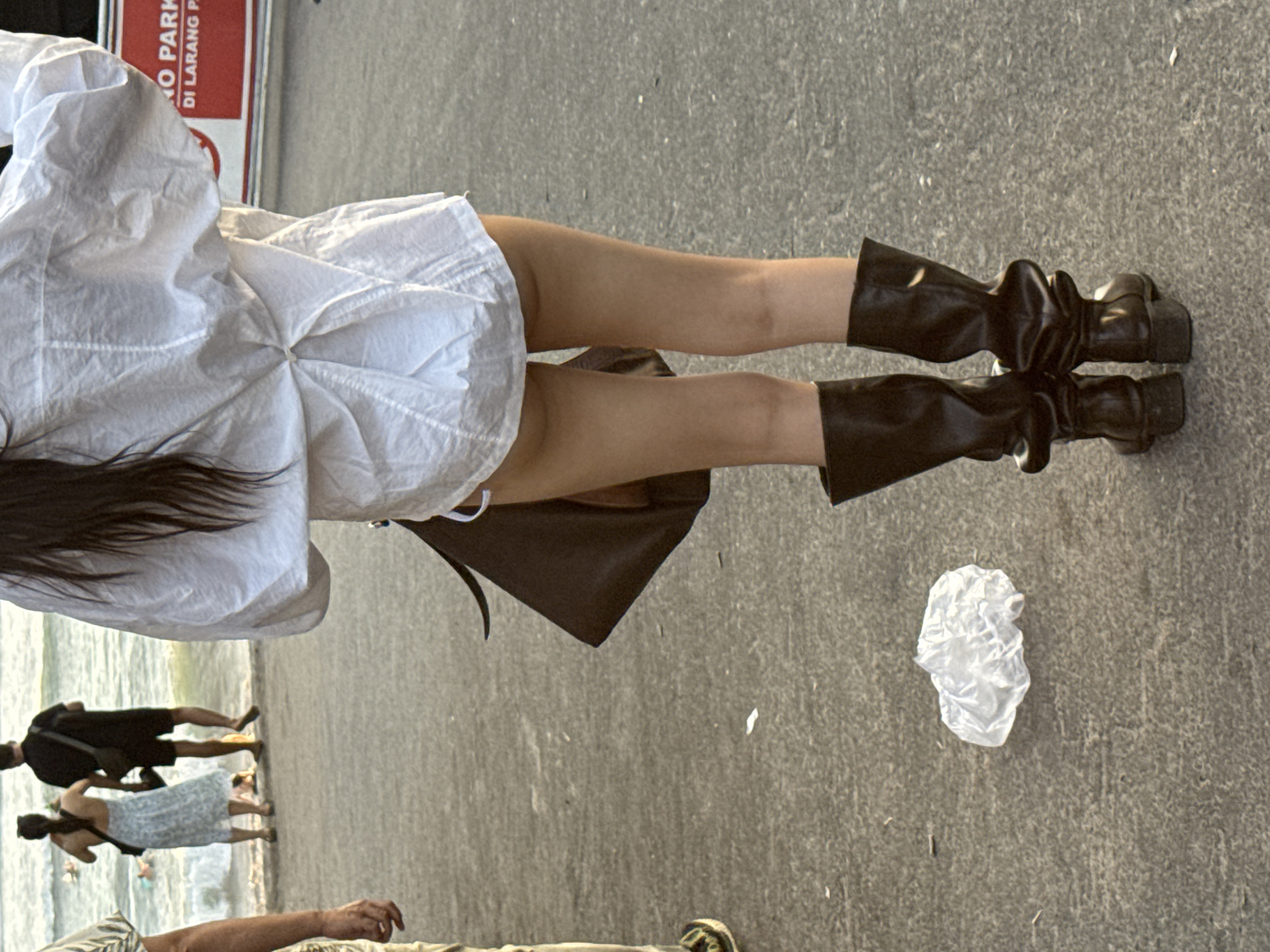 View from the ground of a person wearing a short white dress and black leather boots. A black purse hangs from the person's shoulder. In the background, a shoreline can be seen, where multiple people are walking by the water. A crumpled plastic bag sits on the ground. The scene is located in North Kuta, Indonesia.