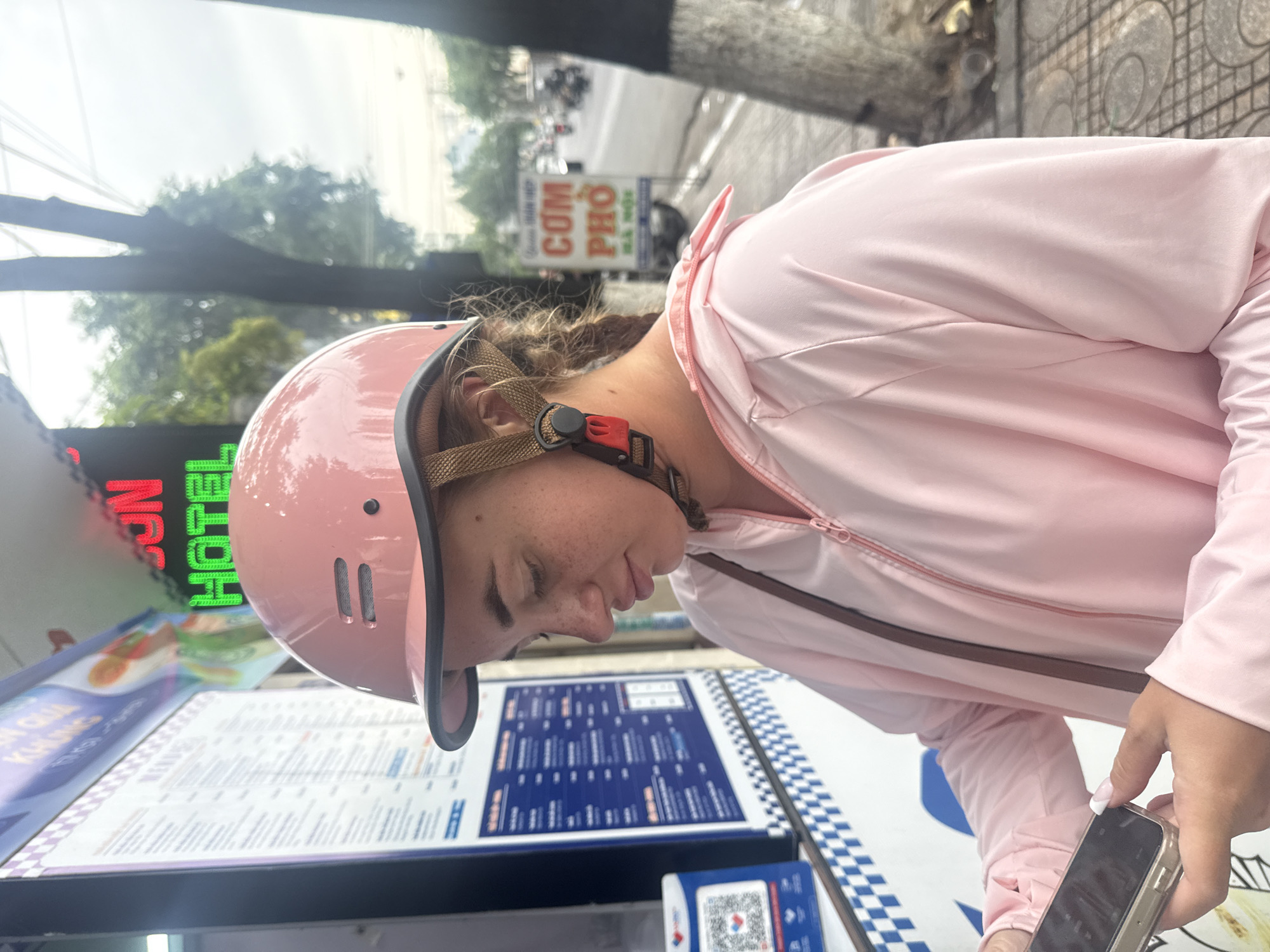 A young Caucasian woman wearing a pink helmet and a pink zip-up jacket is standing on a sidewalk in Nha Trang, Vietnam, looking at her smartphone. She has freckles on her face. Behind her, a menu board with Vietnamese text is visible, along with a QR code. A neon sign with the words "SUN HOTEL" in red and green letters is also present. Further down the street, there is a sign that reads "CƠM PHỞ" and the blur of traffic, including motorcycles, can be seen. The sidewalk is paved with patterned tiles, and a tree trunk is to the right of the woman. The overall scene suggests an urban street setting.