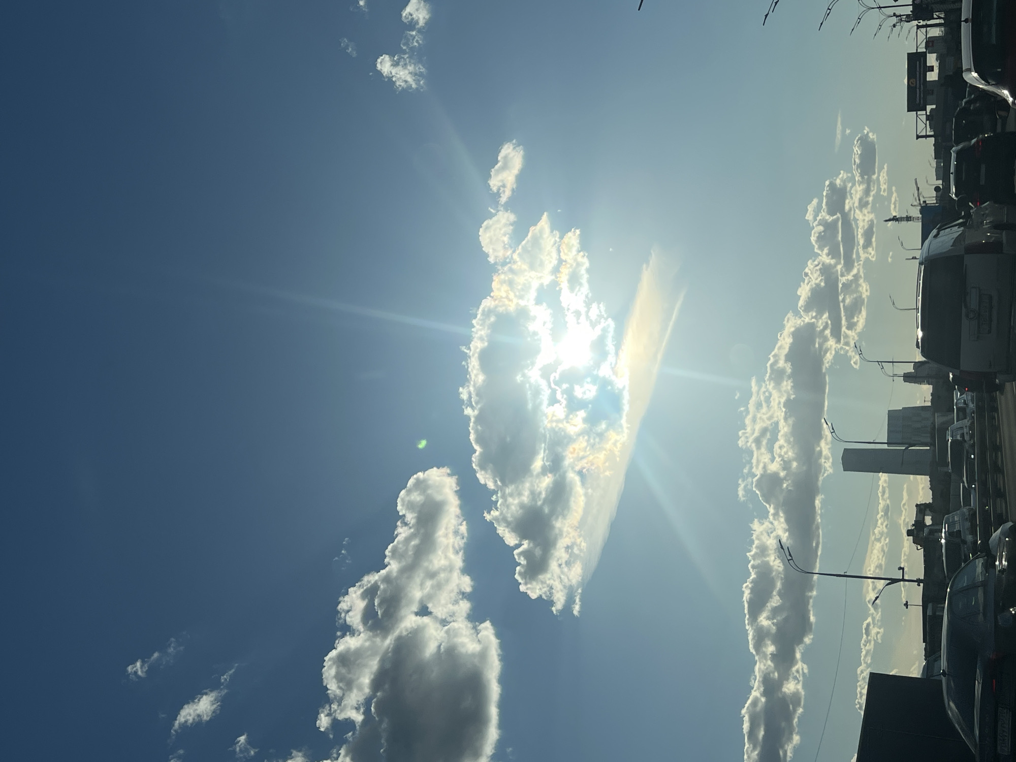 The image depicts a bright, sunny day with a clear blue sky featuring scattered cumulus clouds. Sunlight beams through a larger cloud formation, creating a lens flare effect. Below the sky, a multi-lane highway is visible, with a moderate amount of traffic. Several cars are stopped or moving slowly, suggesting a traffic jam. Streetlights line the highway. In the background, city structures, including tall buildings and construction cranes, are visible, indicating an urban setting. The presence of buildings and the overall cityscape suggest this scene is likely in Moscow, Russia, given the architectural style of the buildings and the known urban development in the city. There are no visible people.