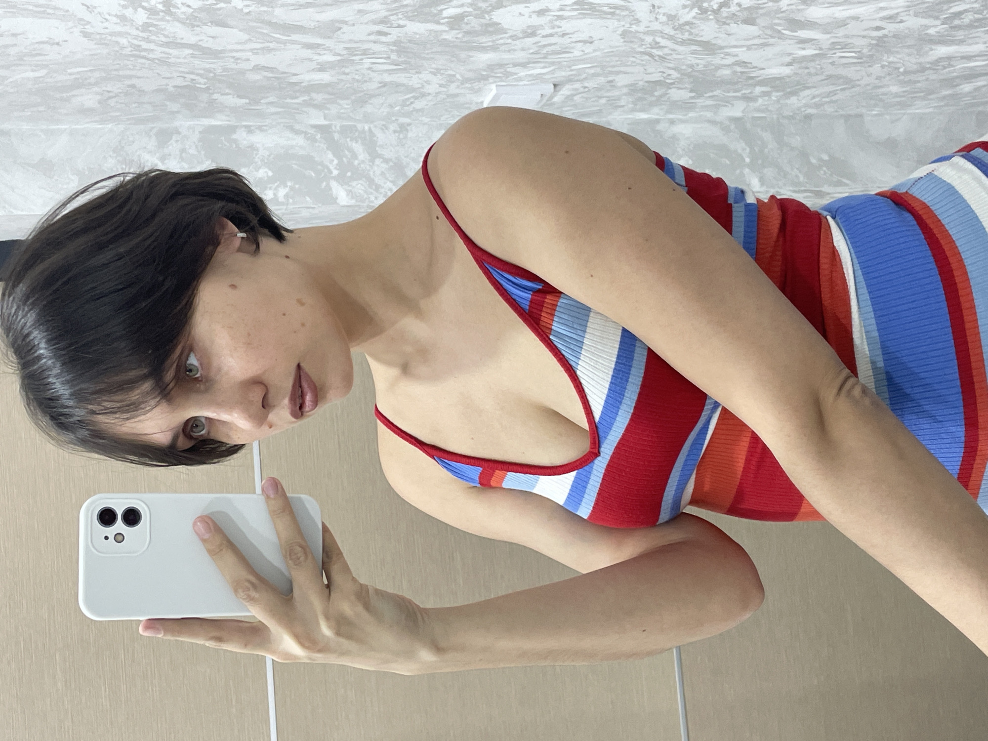 A woman is holding a white smartphone in her left hand, apparently taking a selfie. She has short, dark hair and fair skin, and is wearing a striped, sleeveless top with red, white, and blue stripes. The background is a textured wall with white and gray marble-like pattern above a plain beige wall. The location cannot be determined from the image.