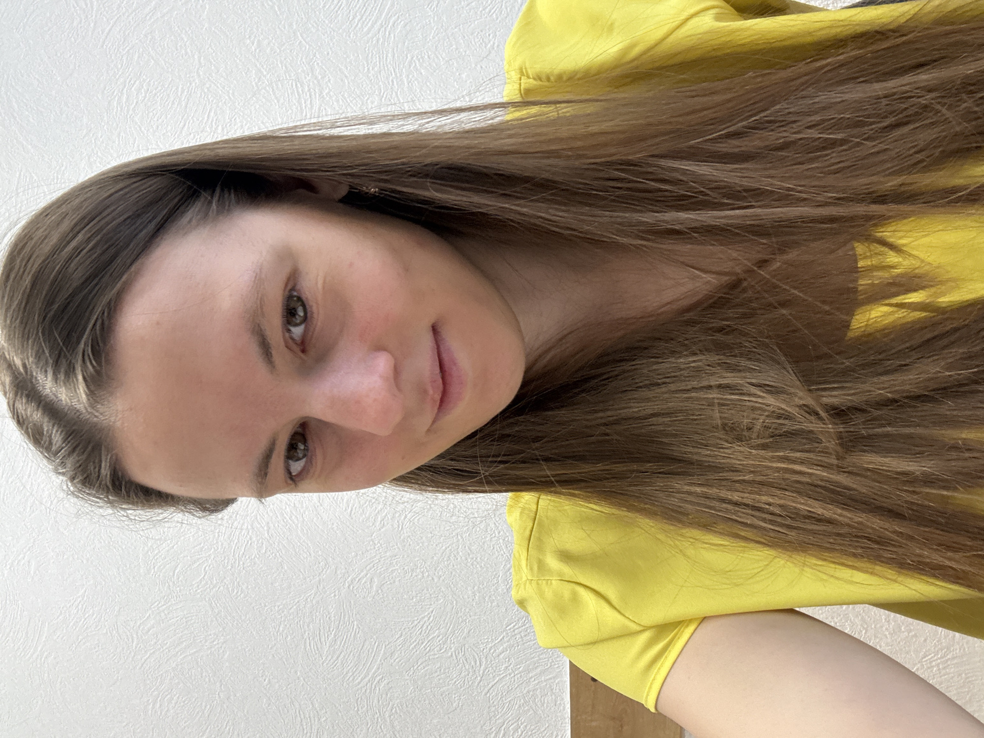 A woman with long, brown hair is facing the camera, wearing a yellow top. She has fair skin and light-colored eyes. The background consists of a textured white wall and a wooden surface. There's no specific information suggesting the location is Chaikovsky, Russia.