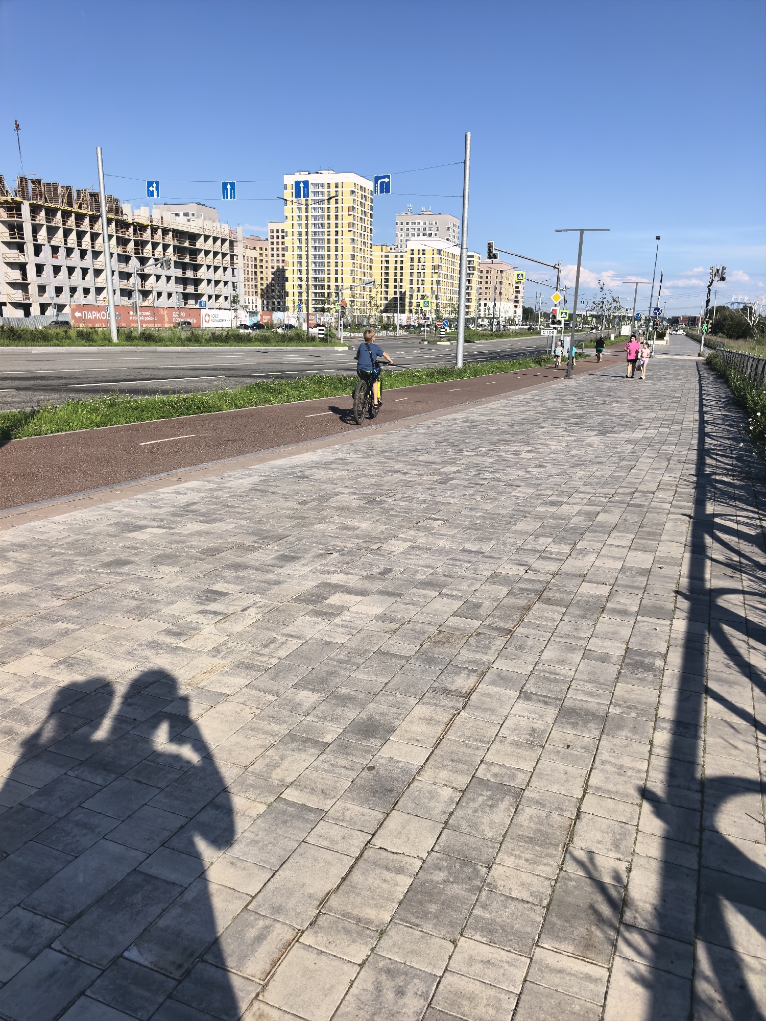 The image captures a sunny day in a city. In the foreground, two shadows of people are cast on a paved walkway. The walkway is made of rectangular grey pavers. To the left of the walkway, a red bike path runs parallel to a road. A young person on a bicycle is riding on the bike path. Further down the walkway, several people are walking. The background features a street with cars, traffic lights, and a construction site with unfinished buildings. Beyond the construction site, modern yellow and white buildings rise against a clear blue sky. The scene suggests an urban environment with ongoing development.