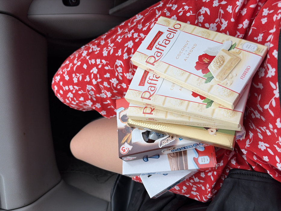 The image displays a close-up, downward-angled view inside a vehicle, centered on the lap of an individual. The left thigh is bare, revealing skin, while the right thigh is covered by a red garment adorned with a white floral pattern. A stack of confectionery products rests on the individual's lap. Specifically, four packages of "Confetteria Raffaello Coconut and Almond" by Ferrero are prominently visible, characterized by white and red branding. Behind these, at least two white-and-red packaged "Kinder" chocolate bars are partially discernible, along with other partially obscured chocolate products, including one with brown packaging and another with gold-colored packaging. The background features generic gray and dark components of a car's interior, consistent with a dashboard or door panel. No specific landmarks or unique environmental features are present to pinpoint an exact location within Budva, Montenegro.