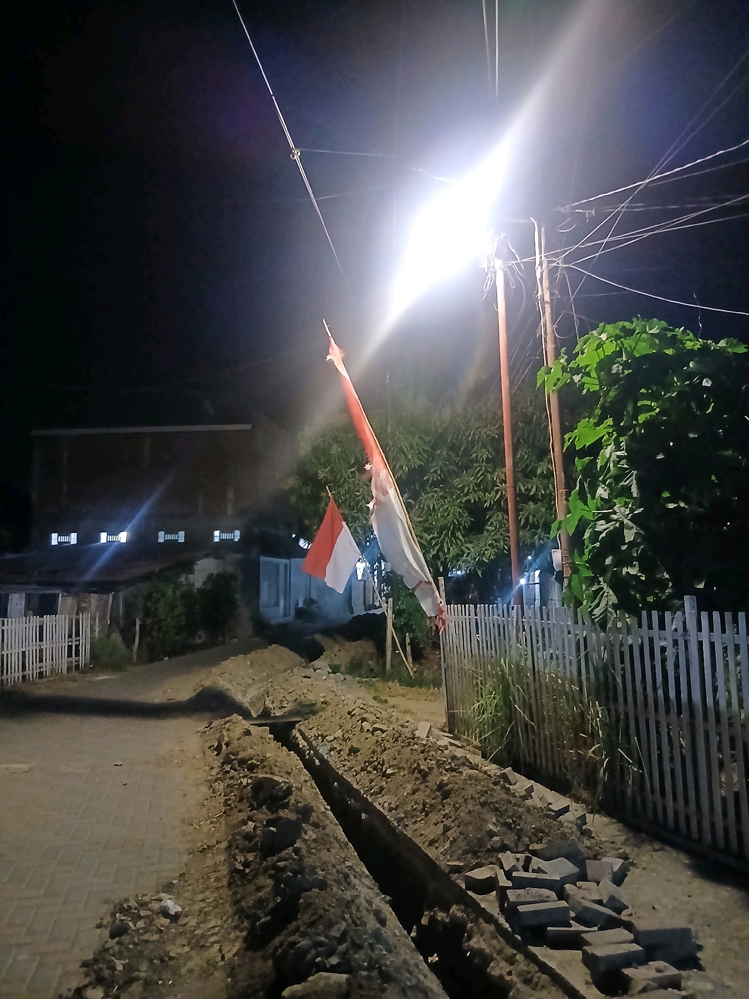 A low-angle, night-time photograph captures a street scene with a bright overhead light illuminating the area. In the foreground, a trench has been dug along a paved street, with piles of dirt and scattered bricks beside it. A white wooden fence runs along the right side of the street, with green foliage visible beyond it. Two Indonesian flags are mounted on a pole, with one flag partially visible in the upper right quadrant of the frame. In the background, a building with multiple windows is visible. The overall impression is one of ongoing construction or repair work in a residential area.