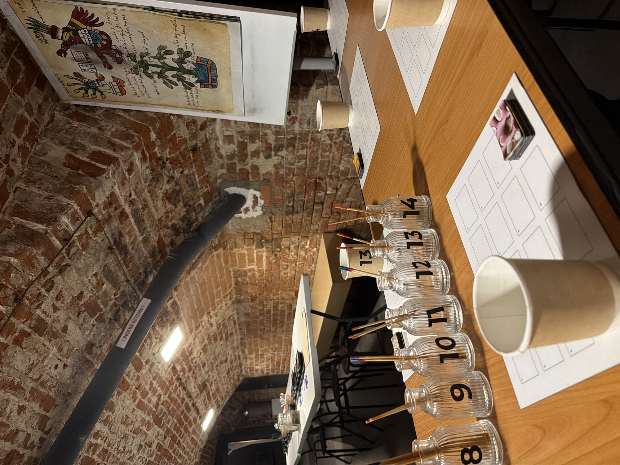 The image depicts a classroom or workshop setting, likely in Nizhny Novgorod, Russia, given the visible signage. The room has a vaulted brick ceiling with exposed pipes, suggesting an older building. On a wooden table in the foreground, there are several glass bottles with numbers 8 through 14, each containing thin sticks with colored tips, presumably for art or craft activities. Also on the table are white paper sheets with grid layouts, brown paper cups, and a small stack of colorful cards. In the background, a long white table is set up with chairs and various containers, possibly holding art supplies. A large poster with intricate drawings and text is displayed on a whiteboard or easel. The lighting is subdued, with illuminated ceiling fixtures. The scene appears to be prepared for a creative session, with an organized arrangement of materials.