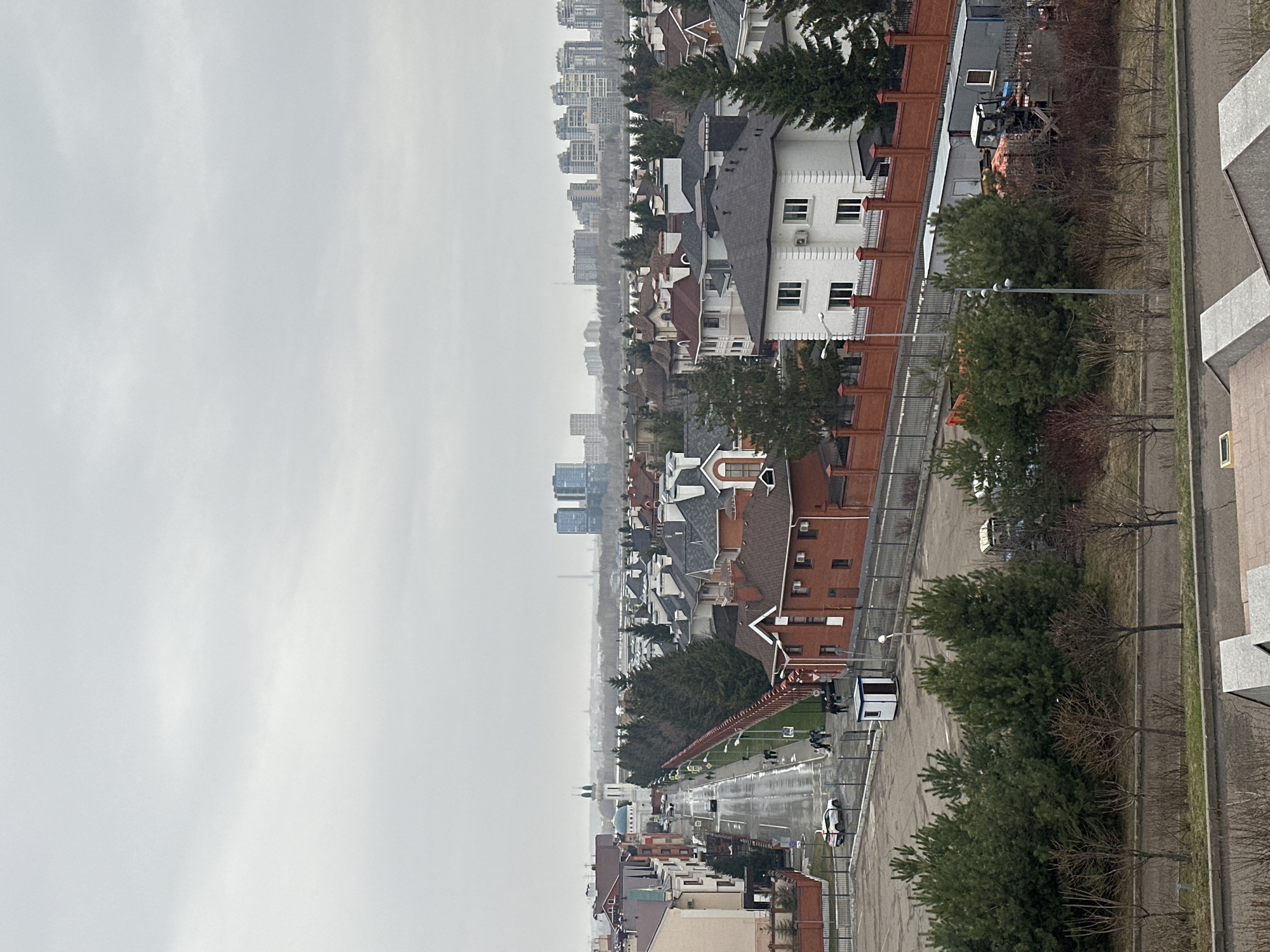 View of Kazan, Russia on a cloudy day, looking down from a height. The scene includes rooftops of buildings, a long, wet street with sparse traffic and pedestrians, trees lining the street, a brick fence, and high-rise buildings in the distance under an overcast sky.