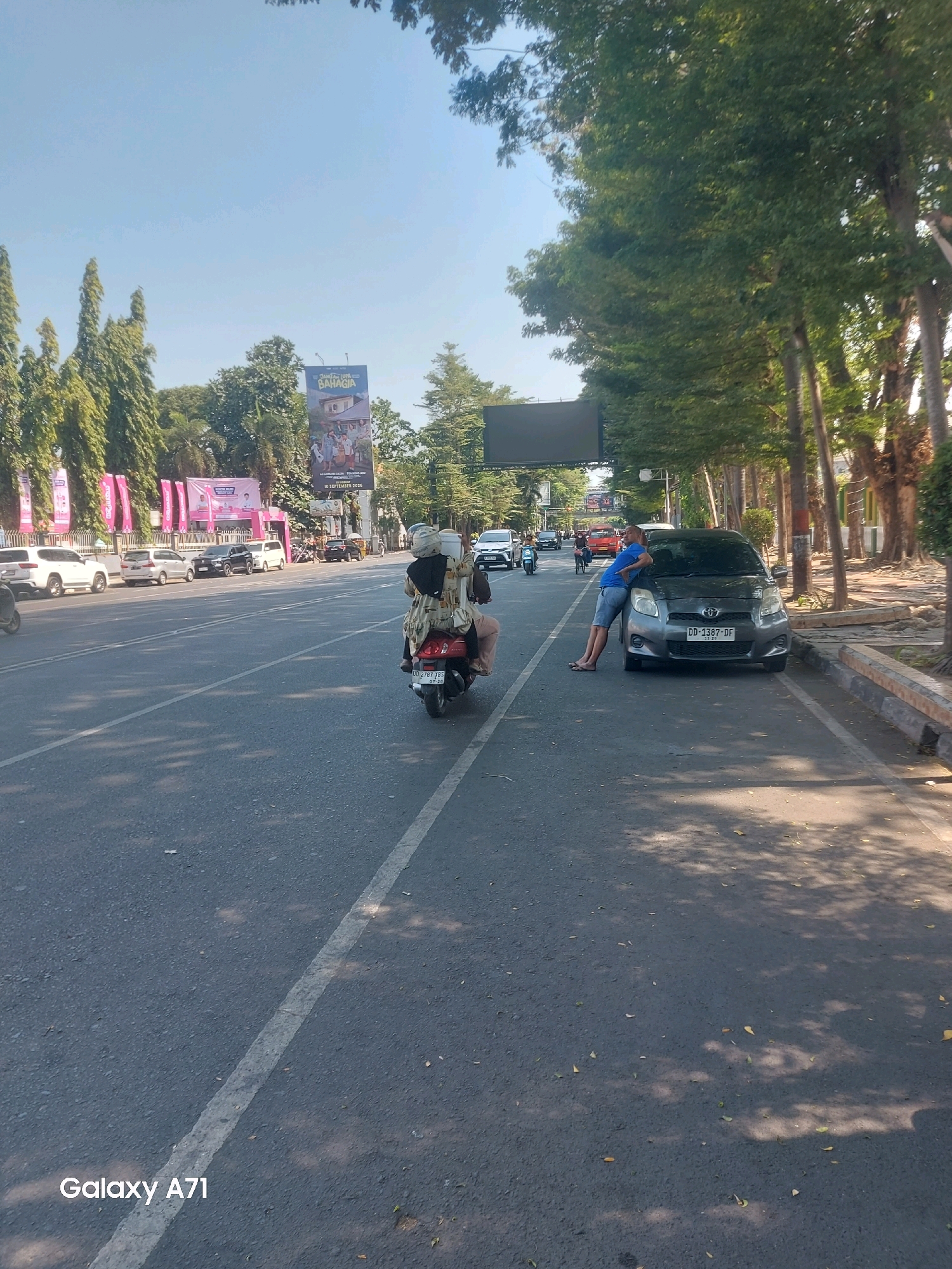 On a sunny day, a dark grey Toyota Yaris with license plate DD 1387 DF is parked on the side of a road in Makassar, Indonesia, with its front bumper resting against a sidewalk. A man wearing a blue t-shirt and denim shorts is leaning against the car, his legs bent. The road is paved with asphalt and has a white dividing line. A scooter with two people on it, one driving and one passenger wearing a headscarf, passes by the car. In the background, there are trees, billboards, and other vehicles on the road. One billboard reads "BAHAGIA" and displays an image of a family. Another billboard is dark and rectangular, possibly a digital screen. The area appears to be a street in a city, with buildings and trees lining the sides of the road.